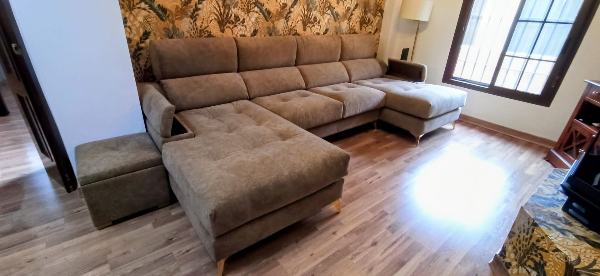 Sala de estar con sofá seccional color beige, otomana y piso de madera cerca de una ventana luminosa.