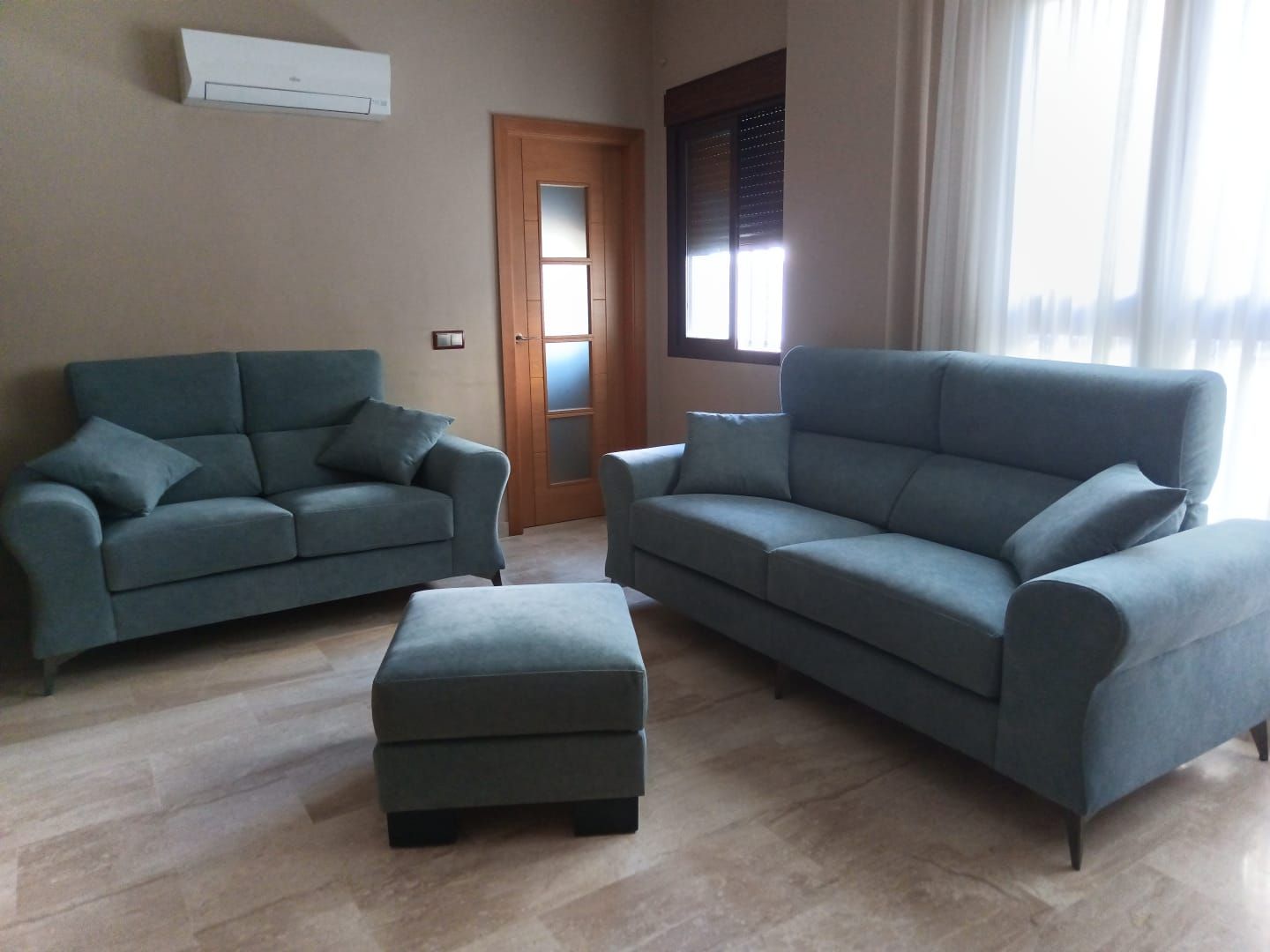 Sala de estar con dos sofás grises, otomana a juego, suelo de madera y ventana con cortinas.