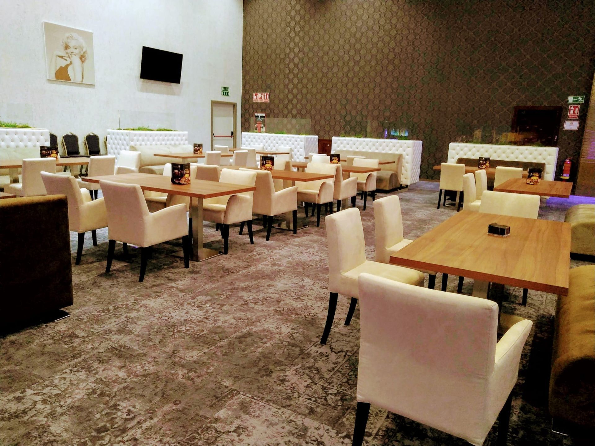 Comedor moderno con sillas beige, mesas de madera y suelo alfombrado, casi vacío.