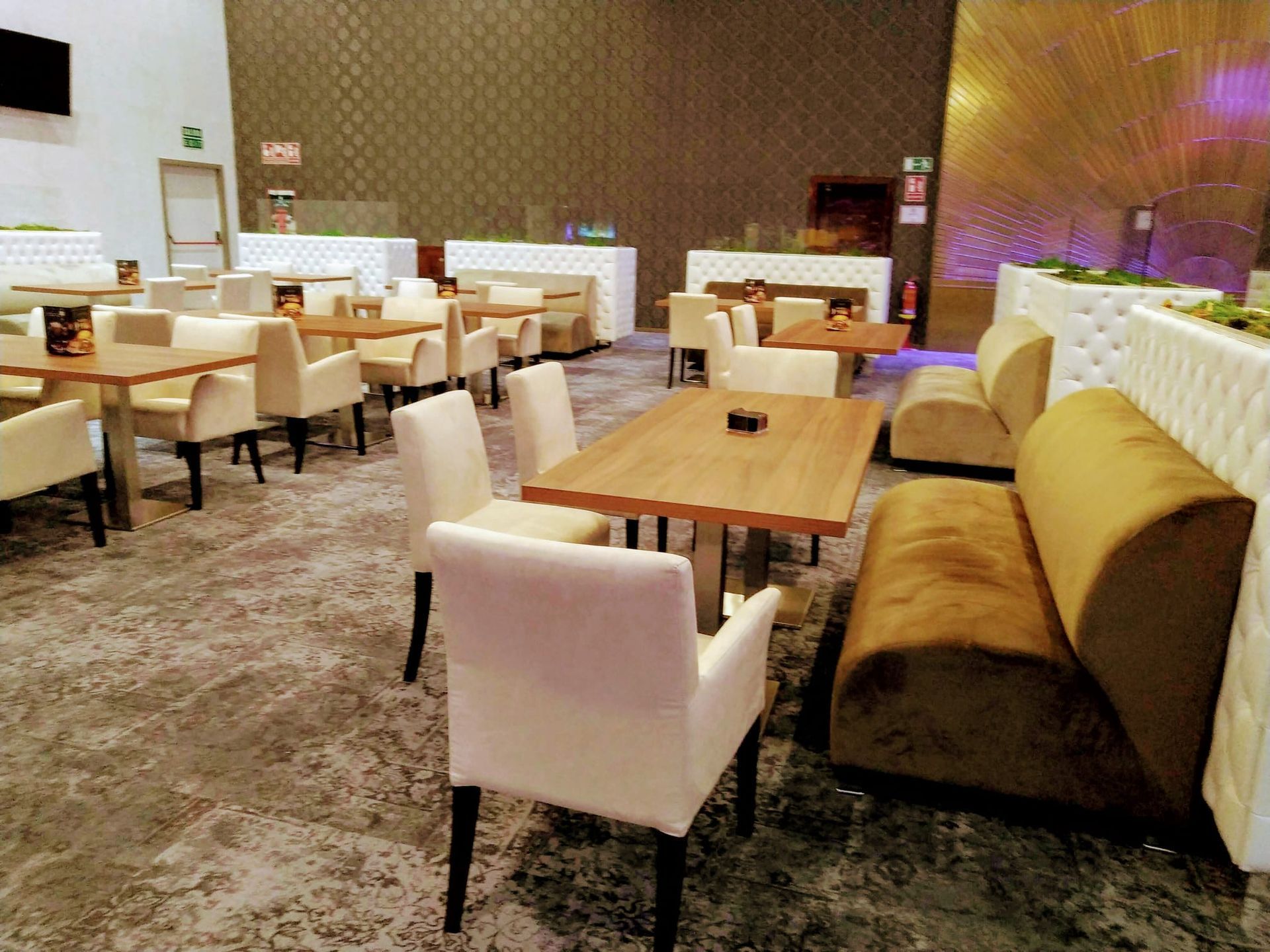 Restaurante moderno y vacío con sillas beige, mesas de madera y un reservado blanco capitoné contra una pared texturizada.