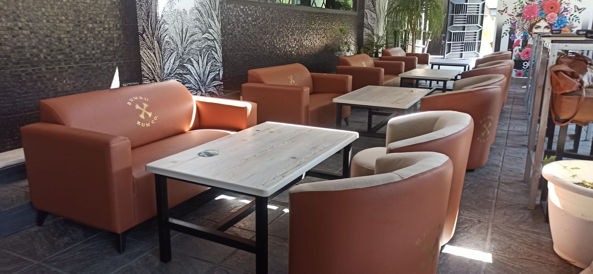 Salón al aire libre con sillones naranjas, sillas beige y mesas en un patio con ladrillos y plantas.