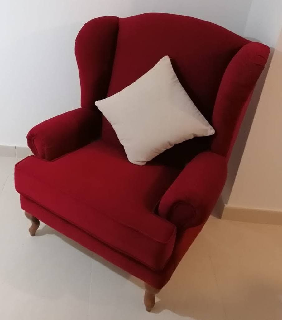 Sillón tapizado en rojo con un cojín blanco en una esquina, contra paredes y suelo claros.