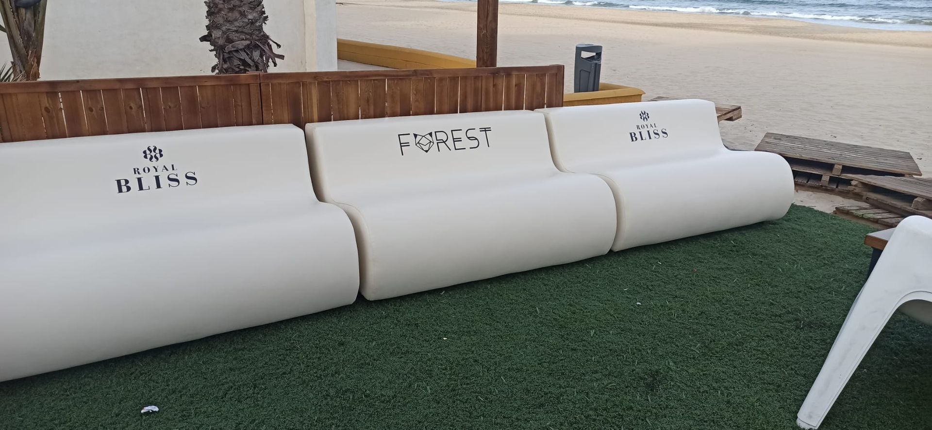 Sofá blanco acolchado para exteriores sobre césped artificial, con respaldos de madera y el océano de fondo.