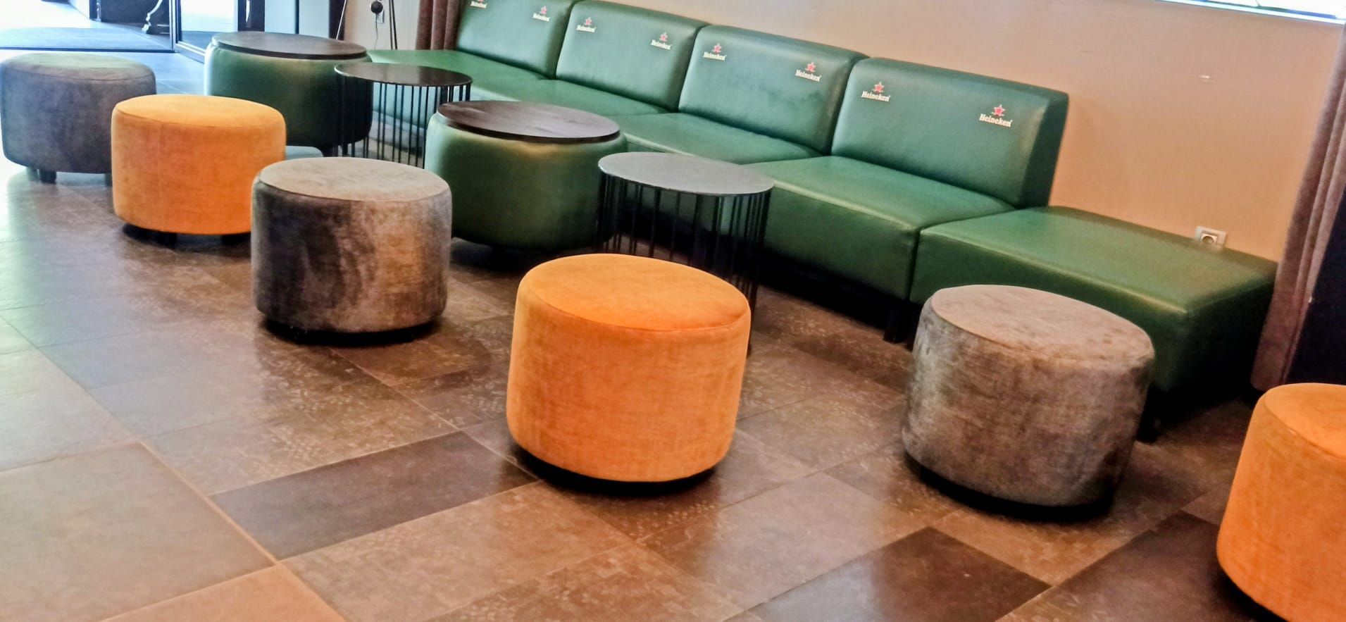 Acogedor salón con sofá modular verde y otomanas redondas naranjas y grises sobre suelo de baldosas.
