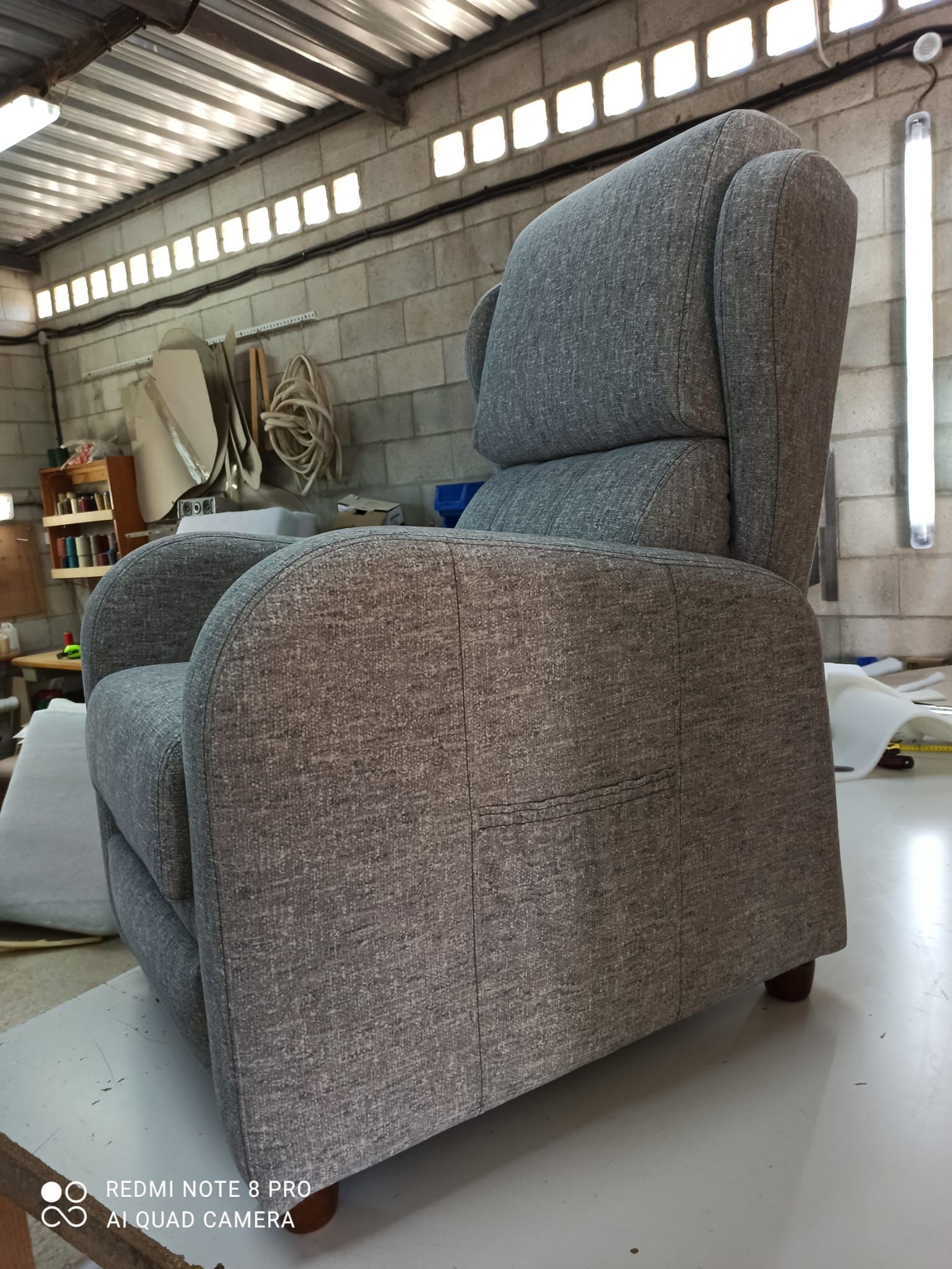 Sillón reclinable tapizado en gris en un taller o garaje con paredes de bloques de hormigón y herramientas al fondo.