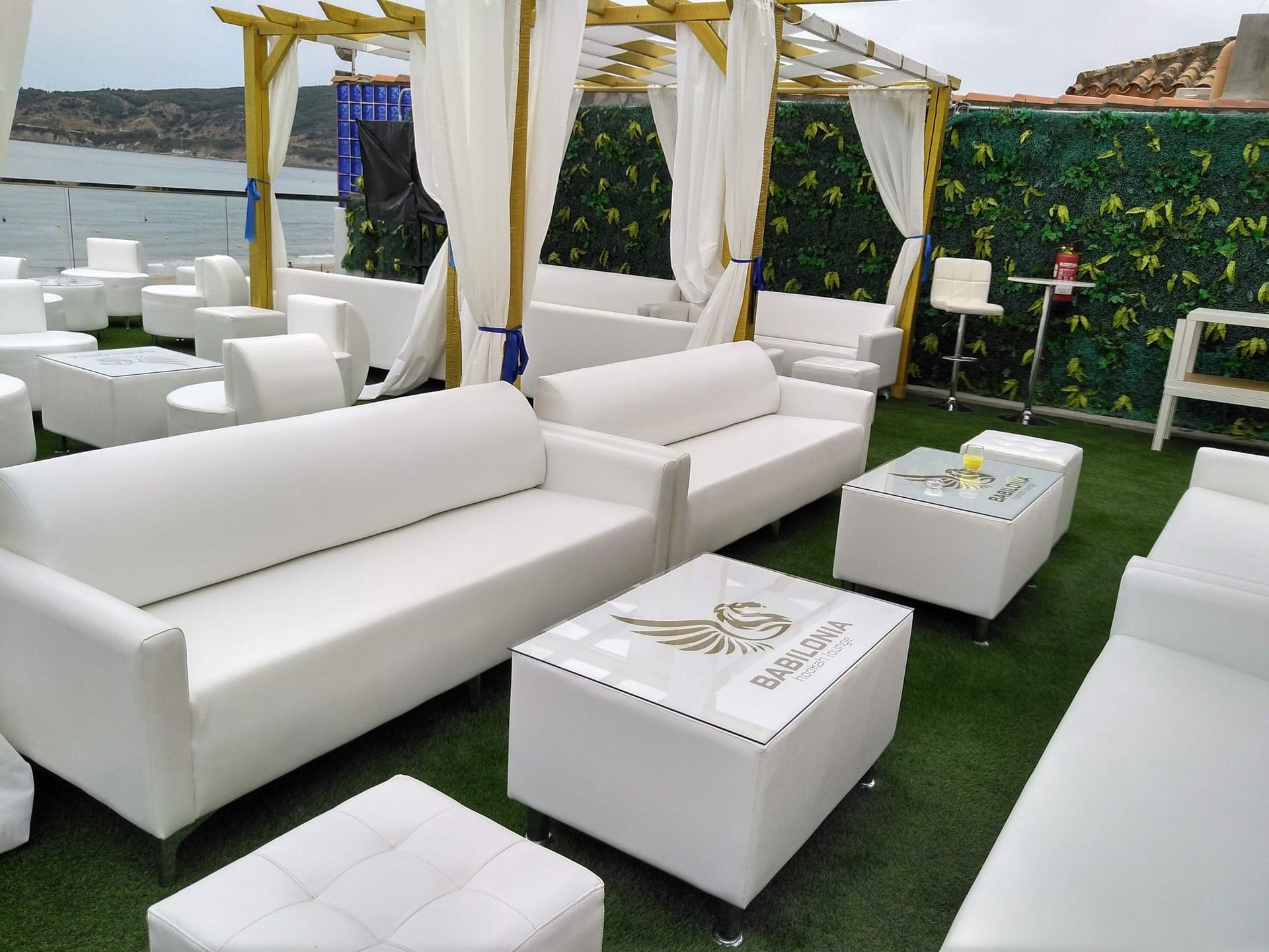 Salón exterior blanco con sofás, otomanas y mesas en una terraza cubierta de césped frente al mar.