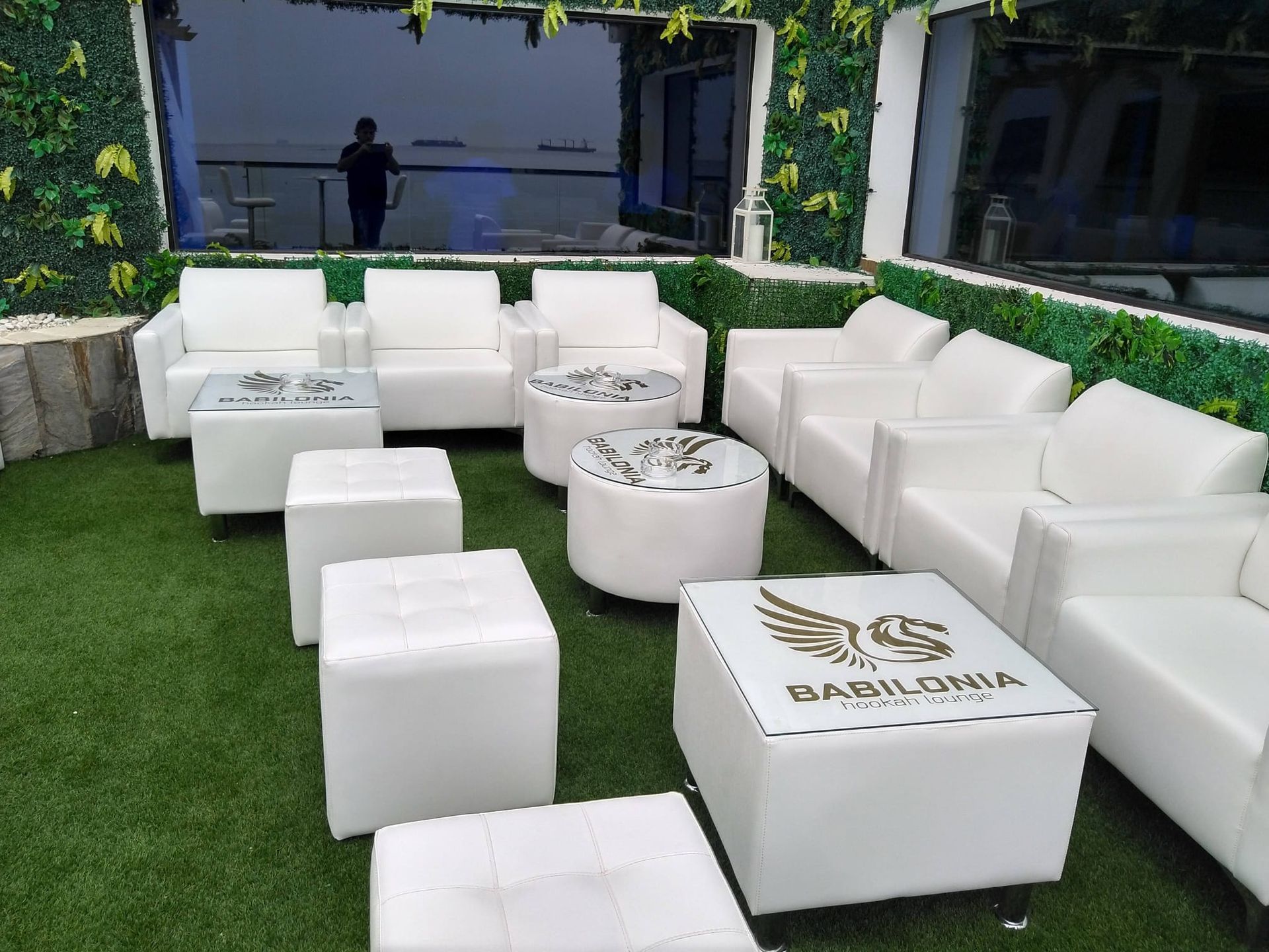 Salón exterior blanco con asientos cúbicos, mesas redondas y paredes cubiertas de hiedra verde