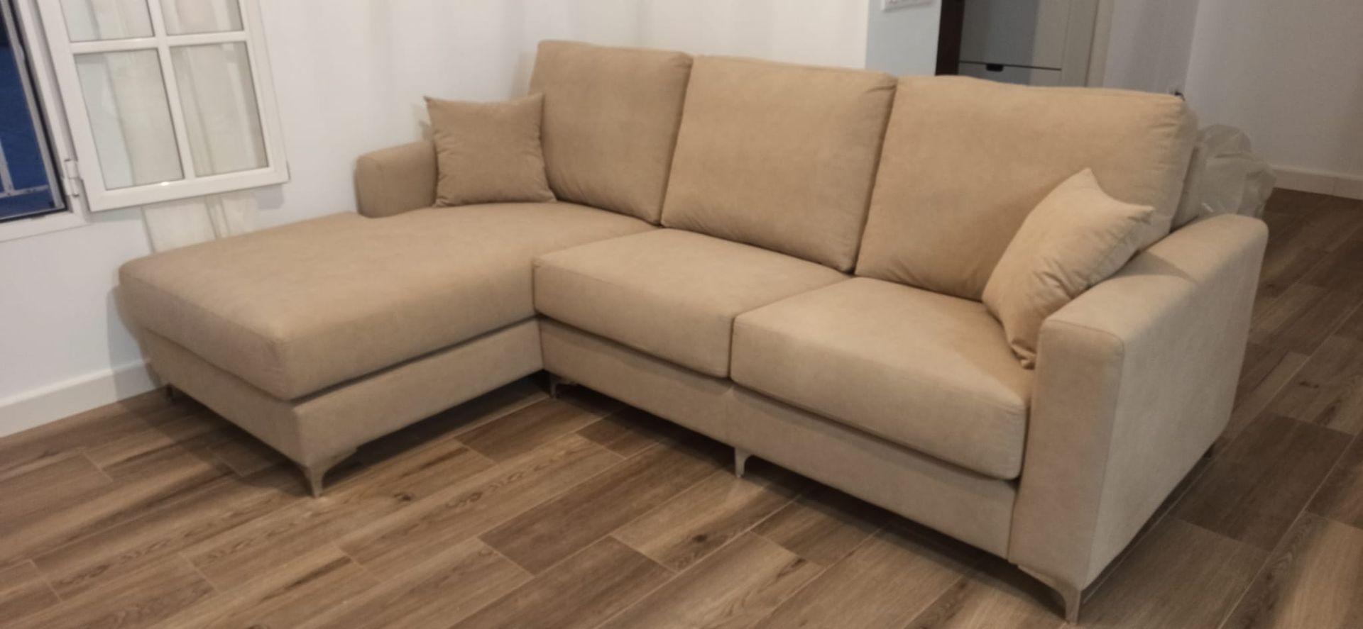 Sofá seccional beige en forma de L en una habitación con suelo de madera y ventana.
