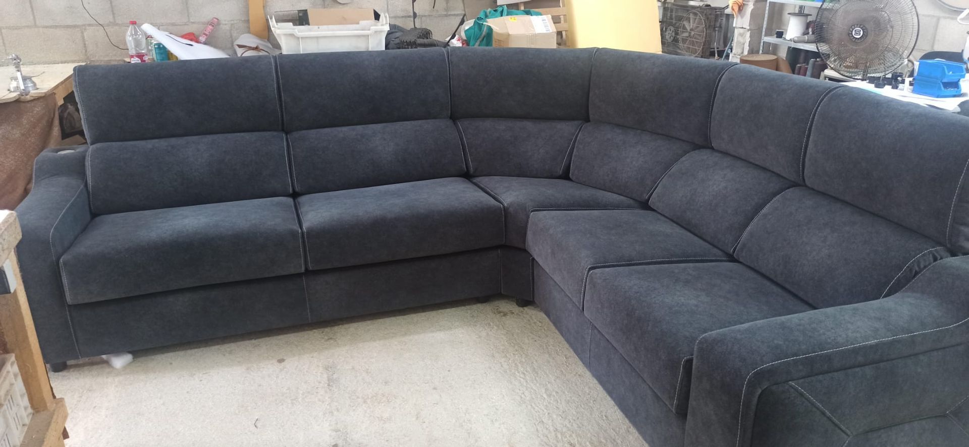 Sofá seccional gris oscuro en una habitación interior, con suelo claro y objetos de taller al fondo.