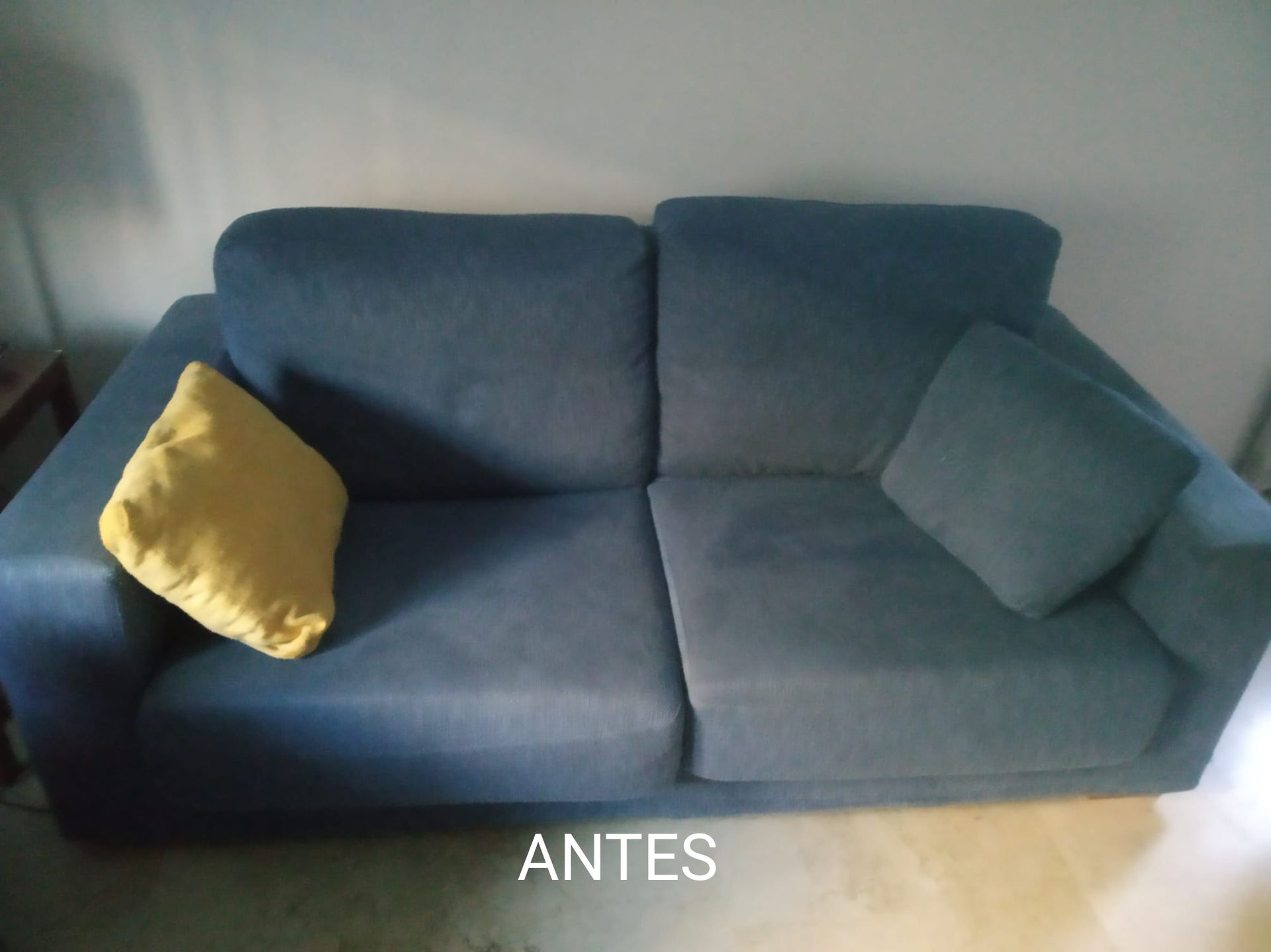 Sofá azul con cojines amarillos y grises contra una pared, con la etiqueta “ANTES” en la parte inferior.