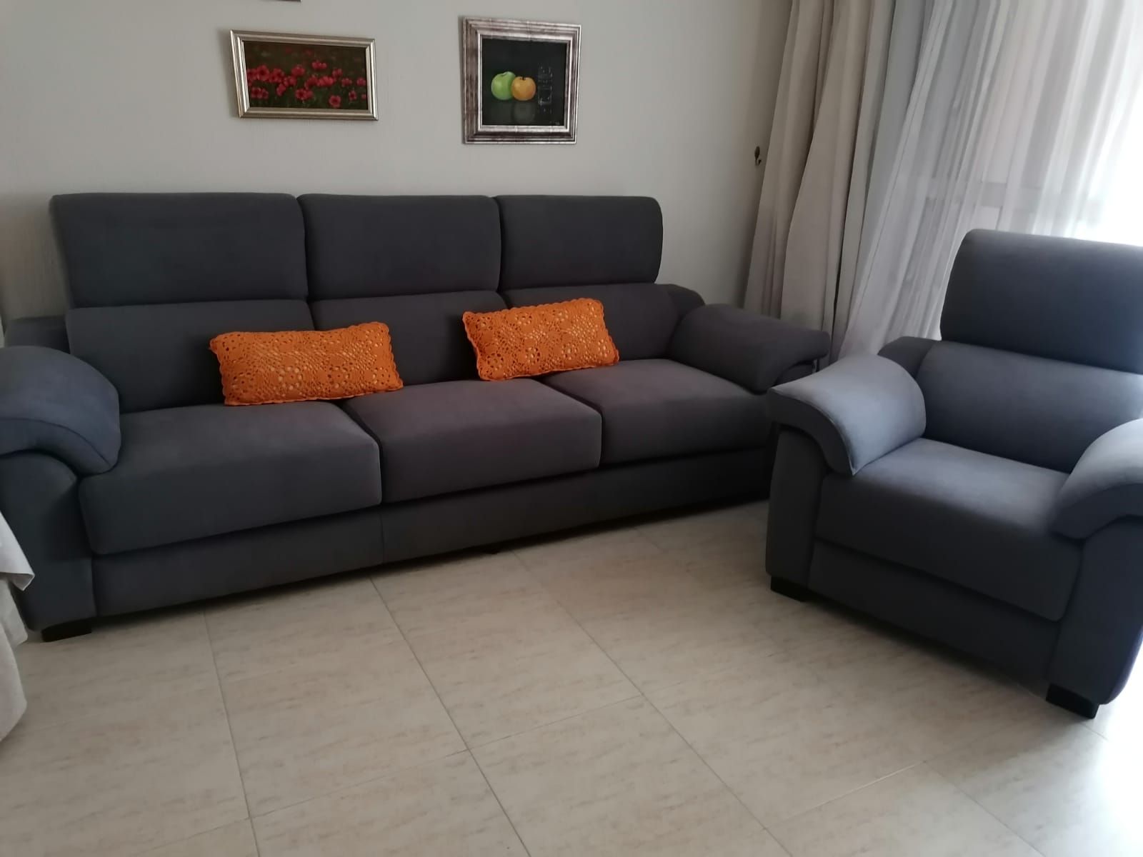 Sofá modular gris y sillón con cojines naranjas en una luminosa sala de estar con suelo de baldosas.