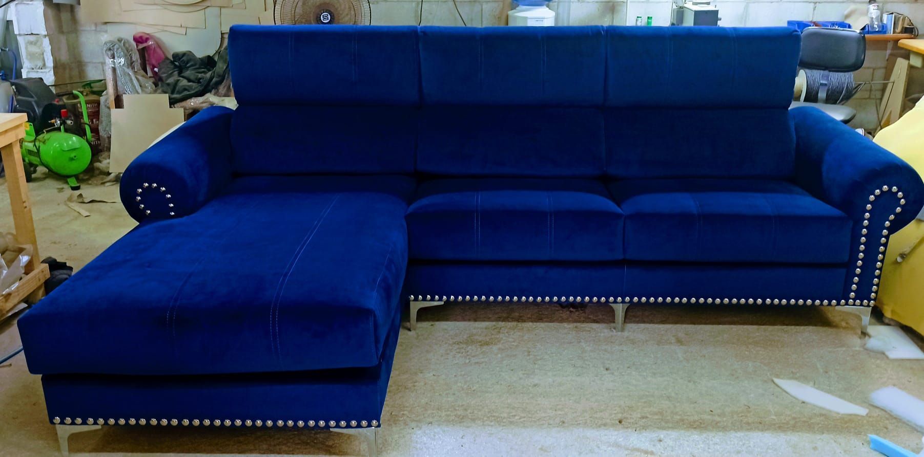 Sofá azul en forma de L en una sala de exposición, con cojines de respaldo capitoné y ribete de tachuelas.