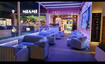 Salón iluminado con luz violeta, con sillones tapizados y la fachada de una tienda de Miami al fondo.
