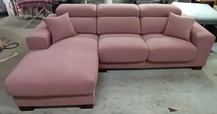 Sofá modular rosa con chaise longue y cojines a juego sobre un suelo de hormigón, expuesto en una sala de exhibición.