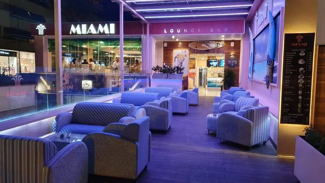 Salón moderno iluminado con luz púrpura, con sofás y mesas, que luce un letrero de Miami y escaparates por la noche.