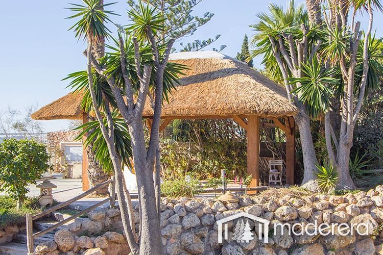 Un cenador de madera con techo de paja está rodeado de árboles y rocas.