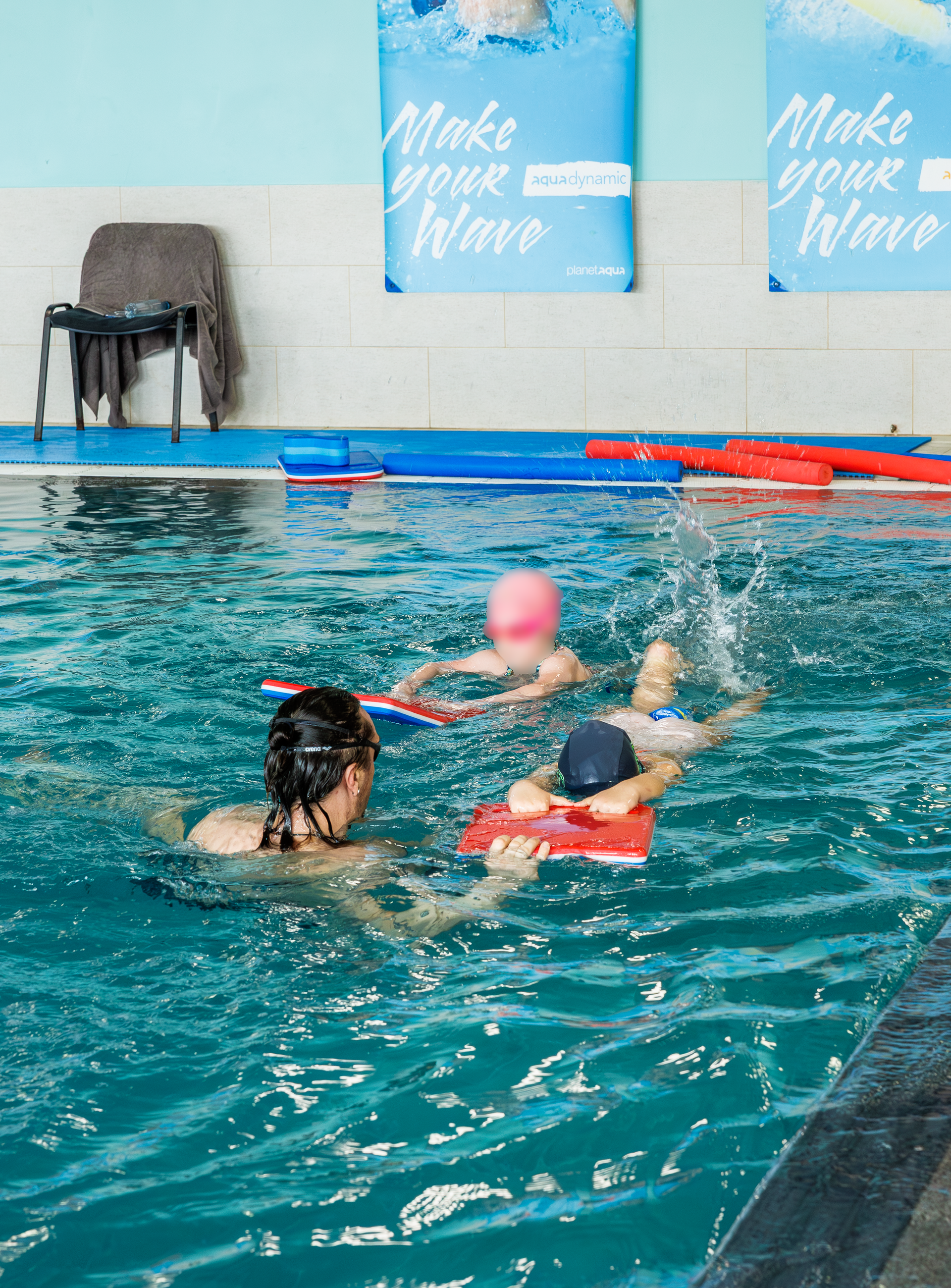 Un moniteur de natation aide deux enfants à s'entraîner avec des planches rouges dans une piscine intérieure.