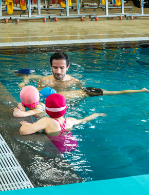 Un enfant souriant, portant des lunettes de natation se tient dans une piscine agrippé à une échelle métallique.
