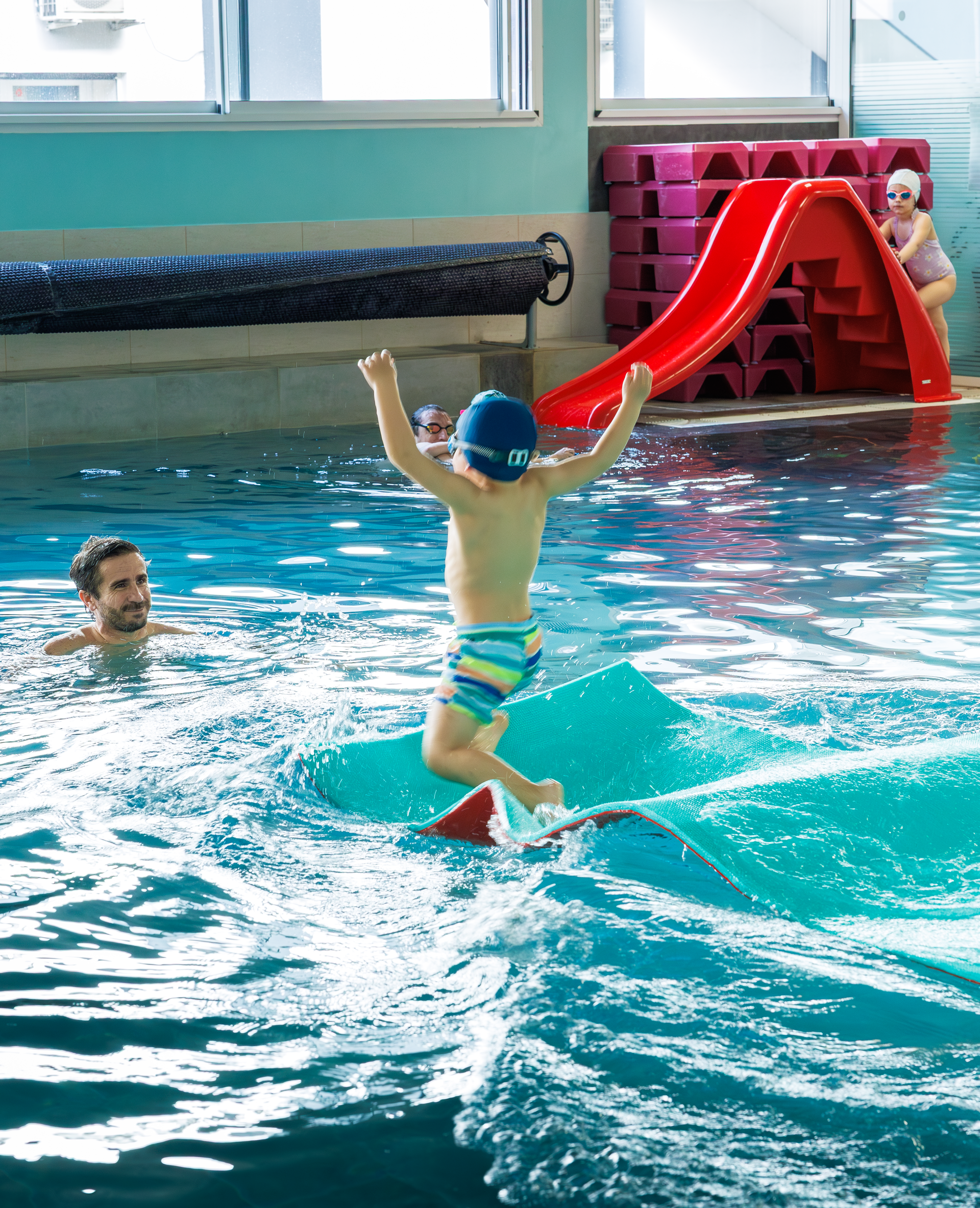 Cinq enfants sourient en tenant des planches de natation rouges au bord d'une piscine.