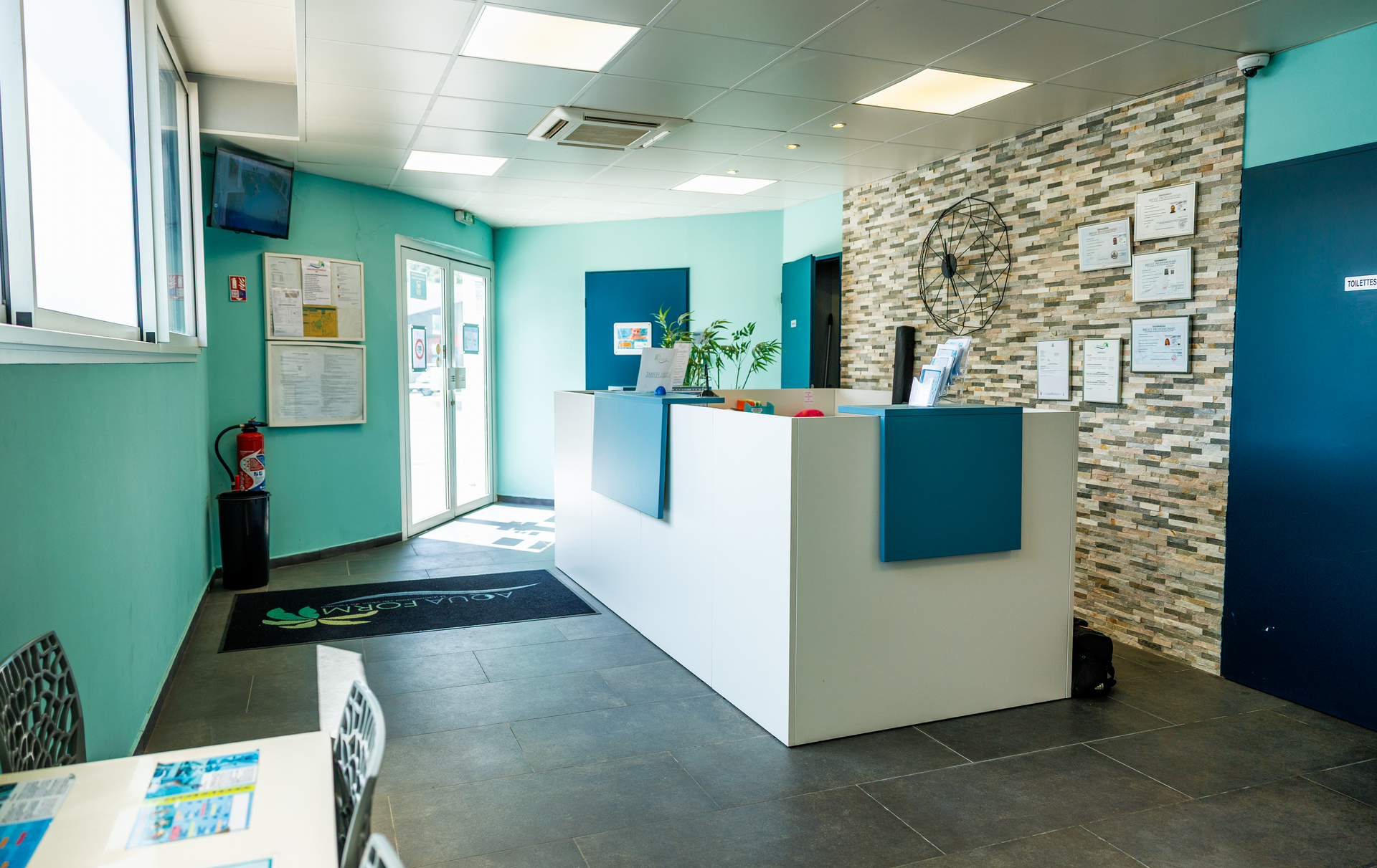 Un espace d'accueil avec un comptoir blanc et bleu, des murs turquoise, un mur du fond orné de pierres et un éclairage vif.
