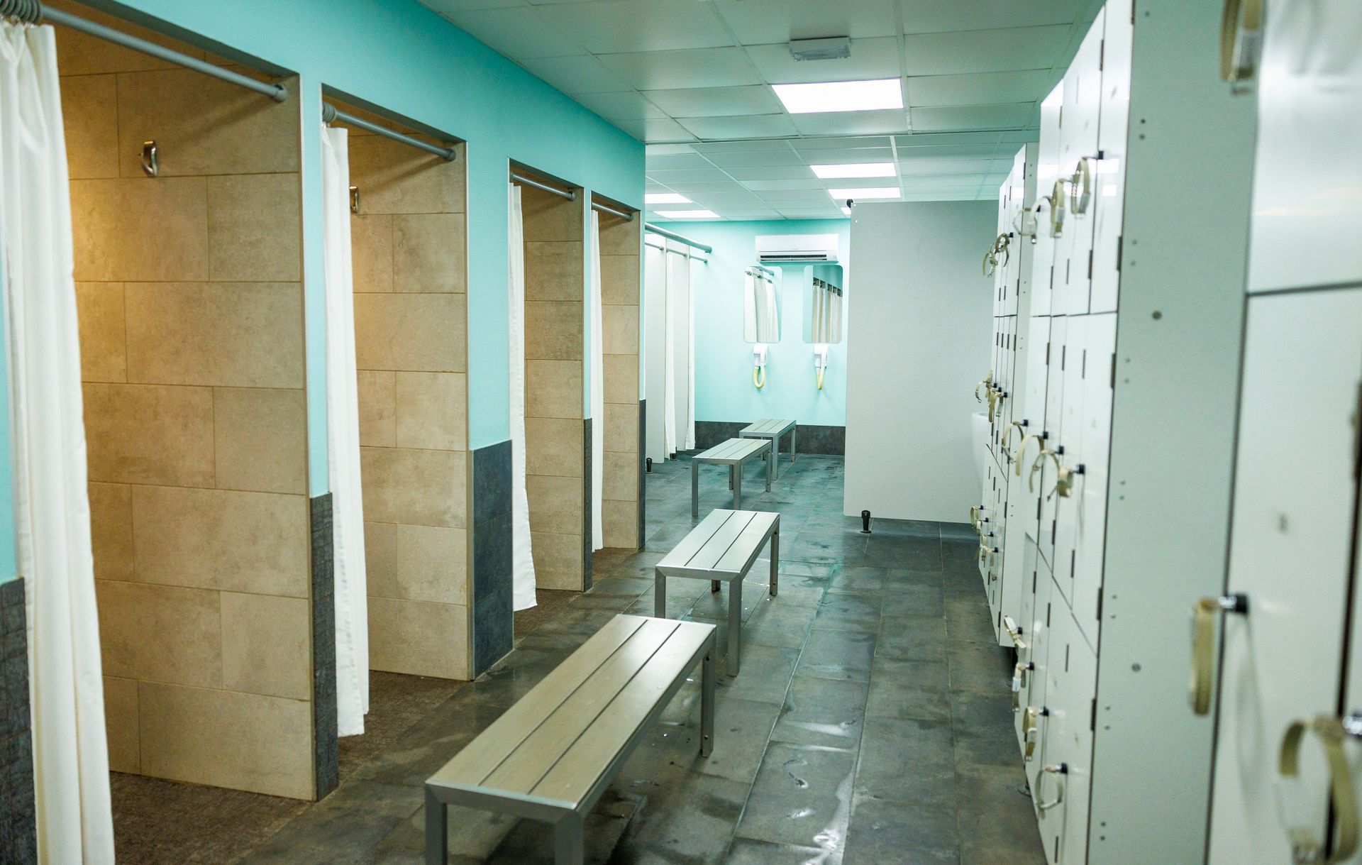 Un vestiaire avec des rangées de casiers blancs, des bancs au centre et des cabines de douche.