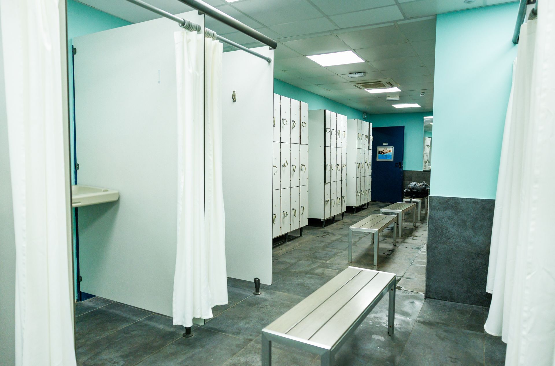 Un vestiaire avec des rideaux blancs pour préserver l'intimité, une rangée de grands casiers blancs et un banc en métal sur un sol carrelé gris.