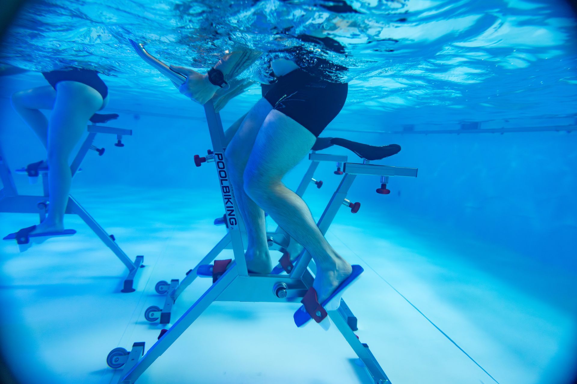 Deux personnes font de l'exercice sur des vélos stationnaires immergés dans une piscine d'un bleu éclatant.