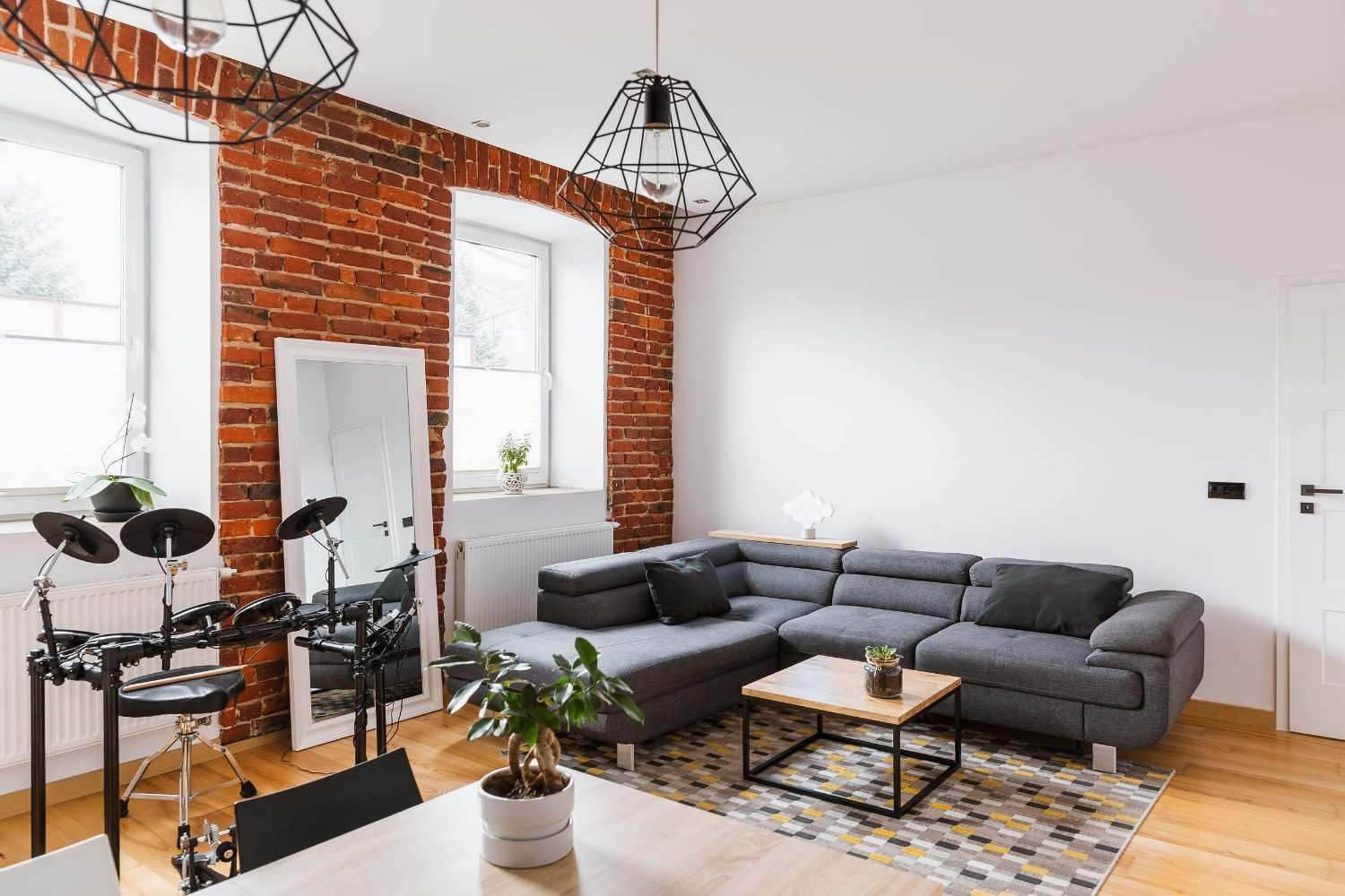 Un salón moderno con paredes de ladrillo visto, un sofá modular gris, una pequeña mesa de centro y una batería electrónica.