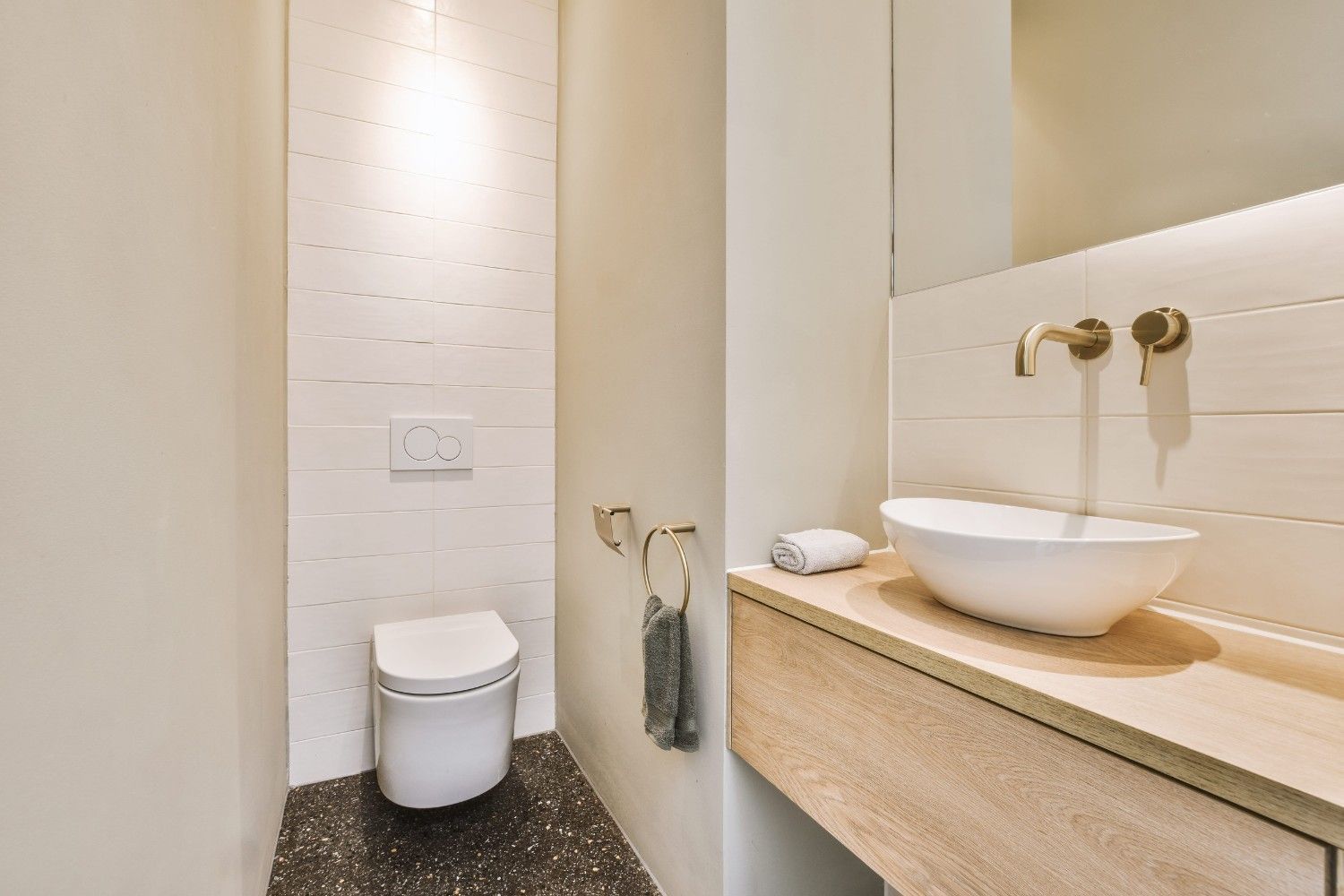 Un baño moderno con inodoro blanco suspendido, tocador flotante de madera con lavabo sobre encimera y grifería dorada.