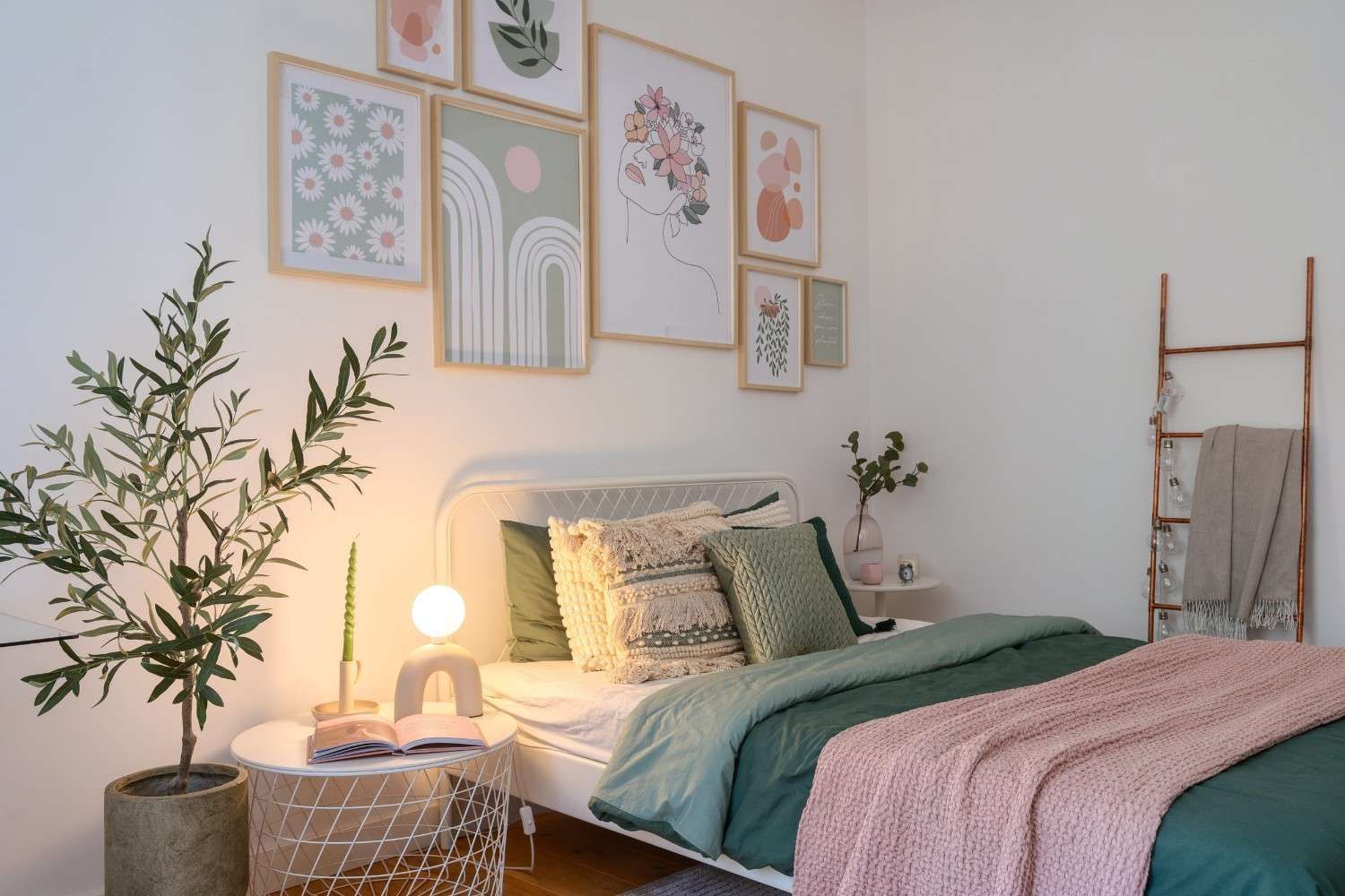 Un dormitorio acogedor con una colcha verde, una manta rosa, cuadros en la pared, una lámpara de noche y un olivo en maceta.