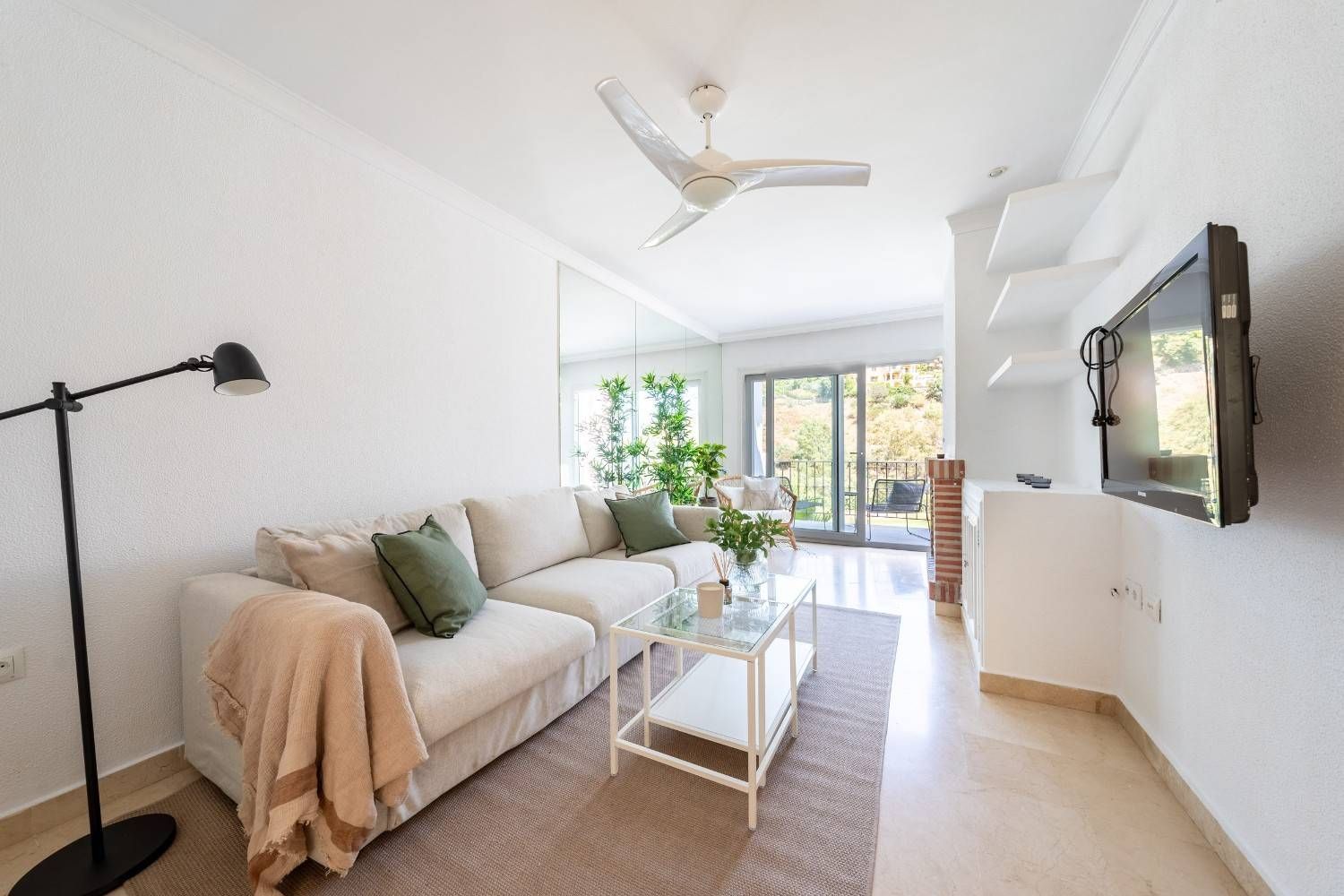 Un salón luminoso y minimalista con un sofá beige, cojines verdes, una mesa de centro de cristal.