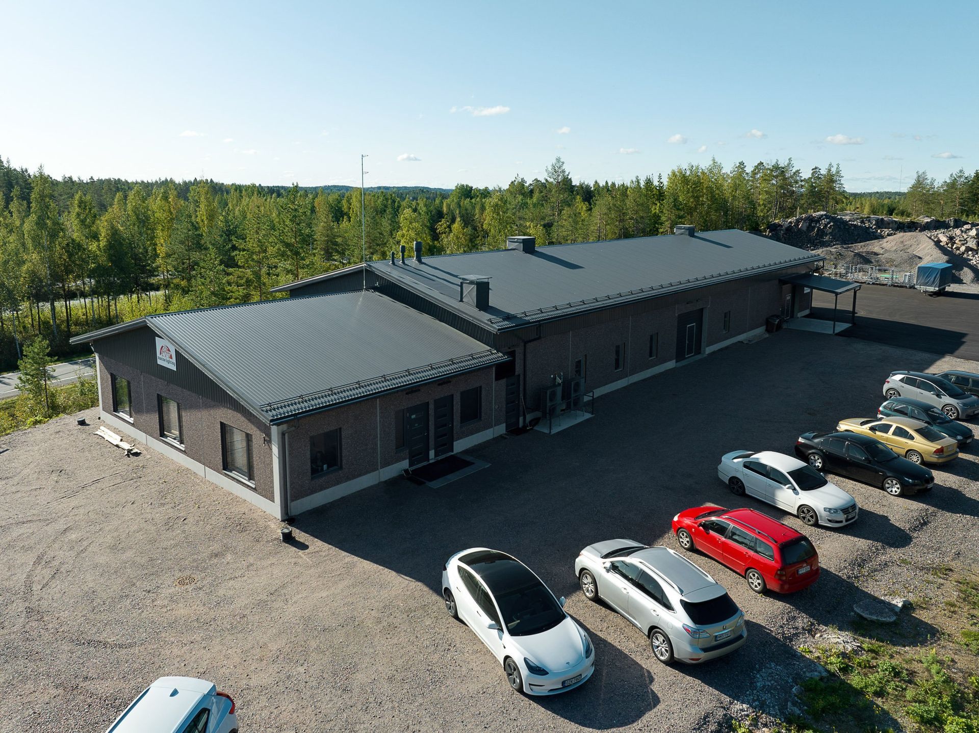 Ilmakuva pitkästä harmaasta rakennuksesta, jonka taustalla on pysäköityjä autoja ja puita.