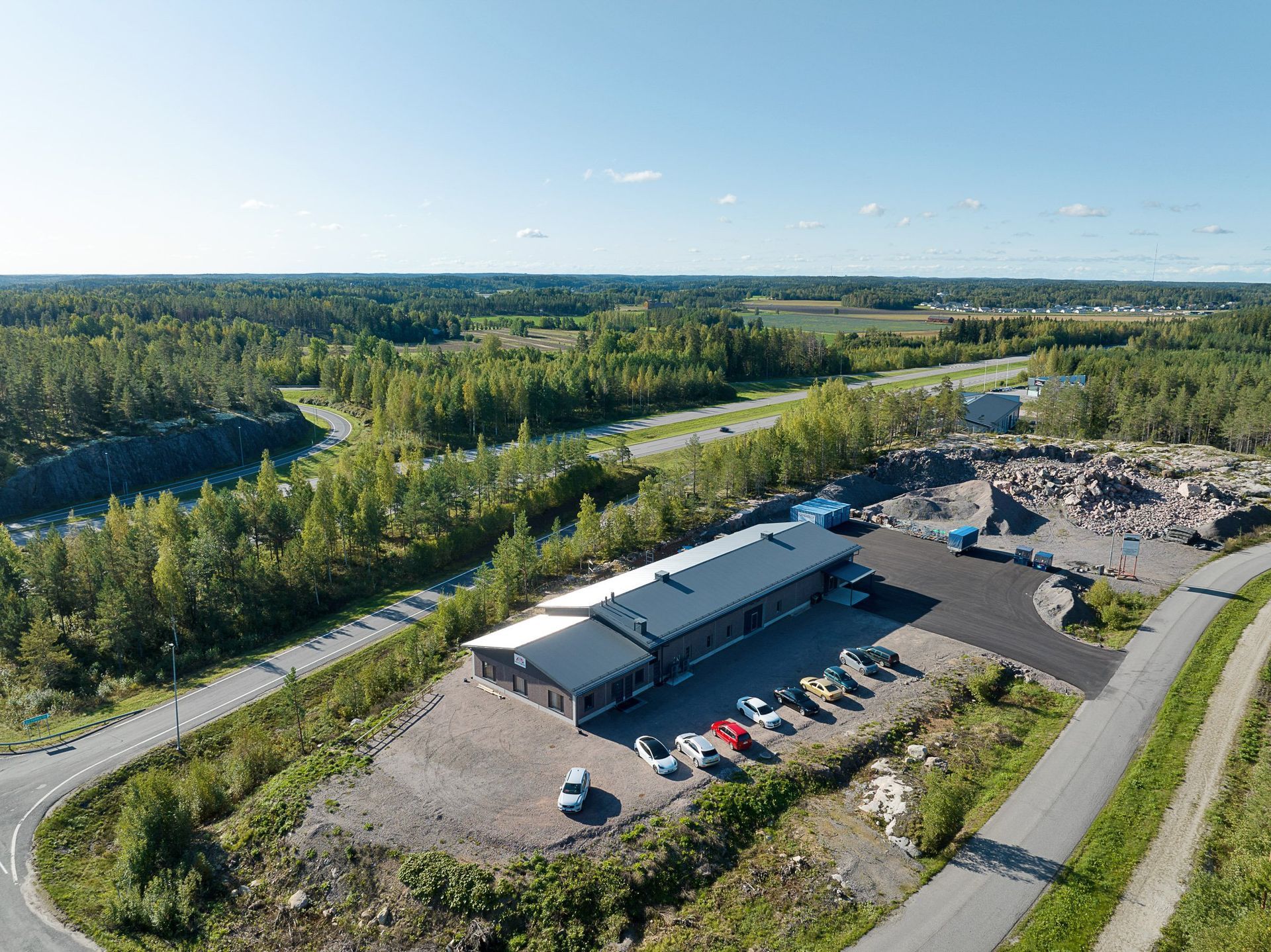 Ilmakuva pienestä teollisuusrakennuksesta, jossa on pysäköityjä autoja, teiden ja tiheän vihreän metsän ympäröimänä.