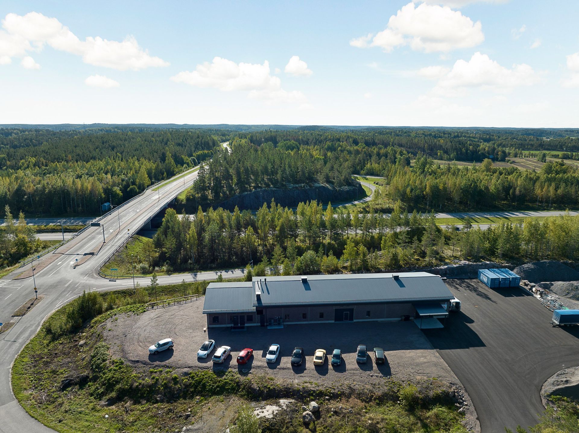 Ilmakuva pitkästä rakennuksesta soratontin ja tien vieressä, tiheän metsän ympäröimänä sinisen taivaan alla.