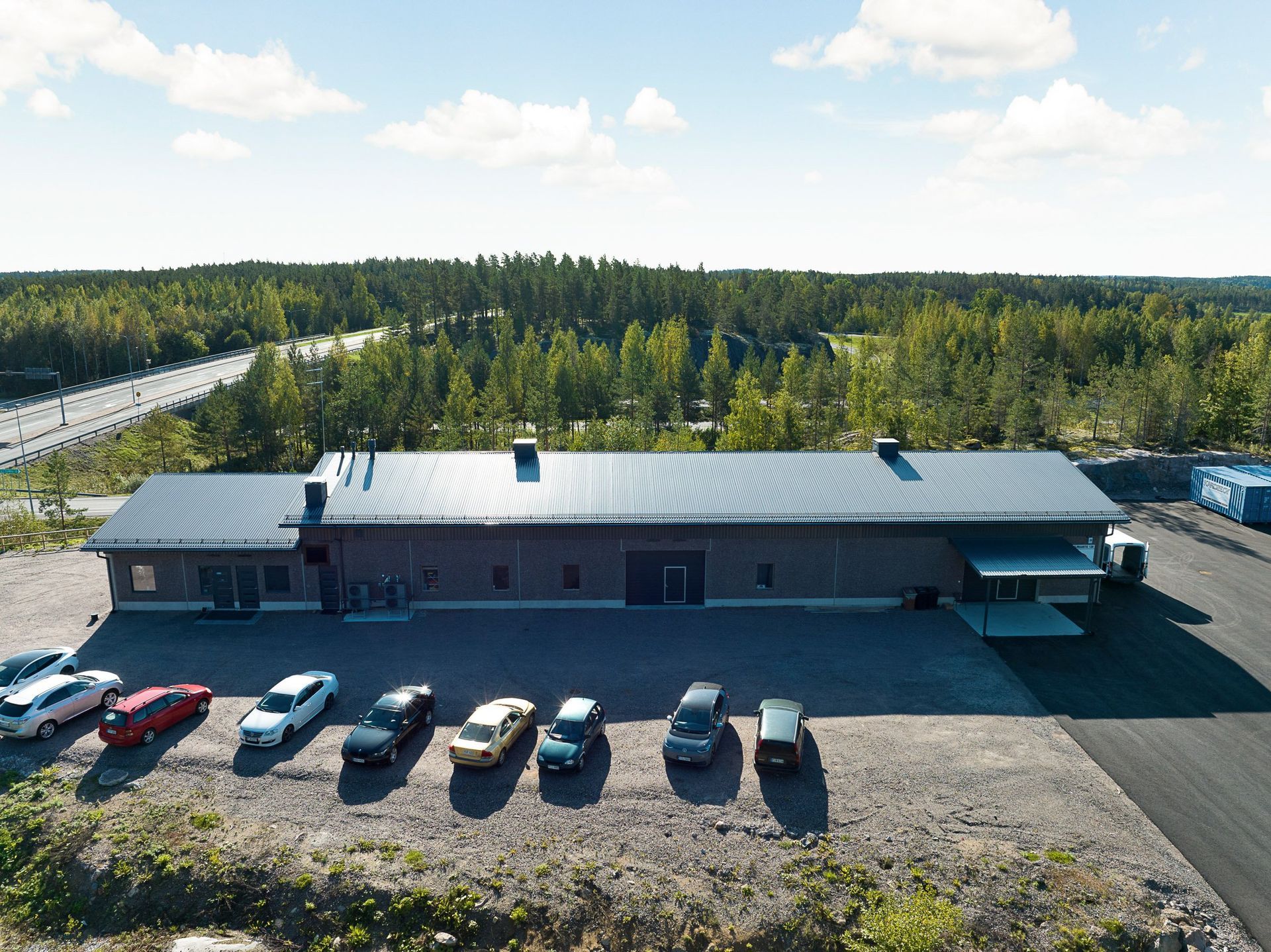 Ilmakuva pitkästä harmaasta rakennuksesta, jonka edessä on pysäköityjä autoja, metsän ja avoimen taivaan ympäröimänä.