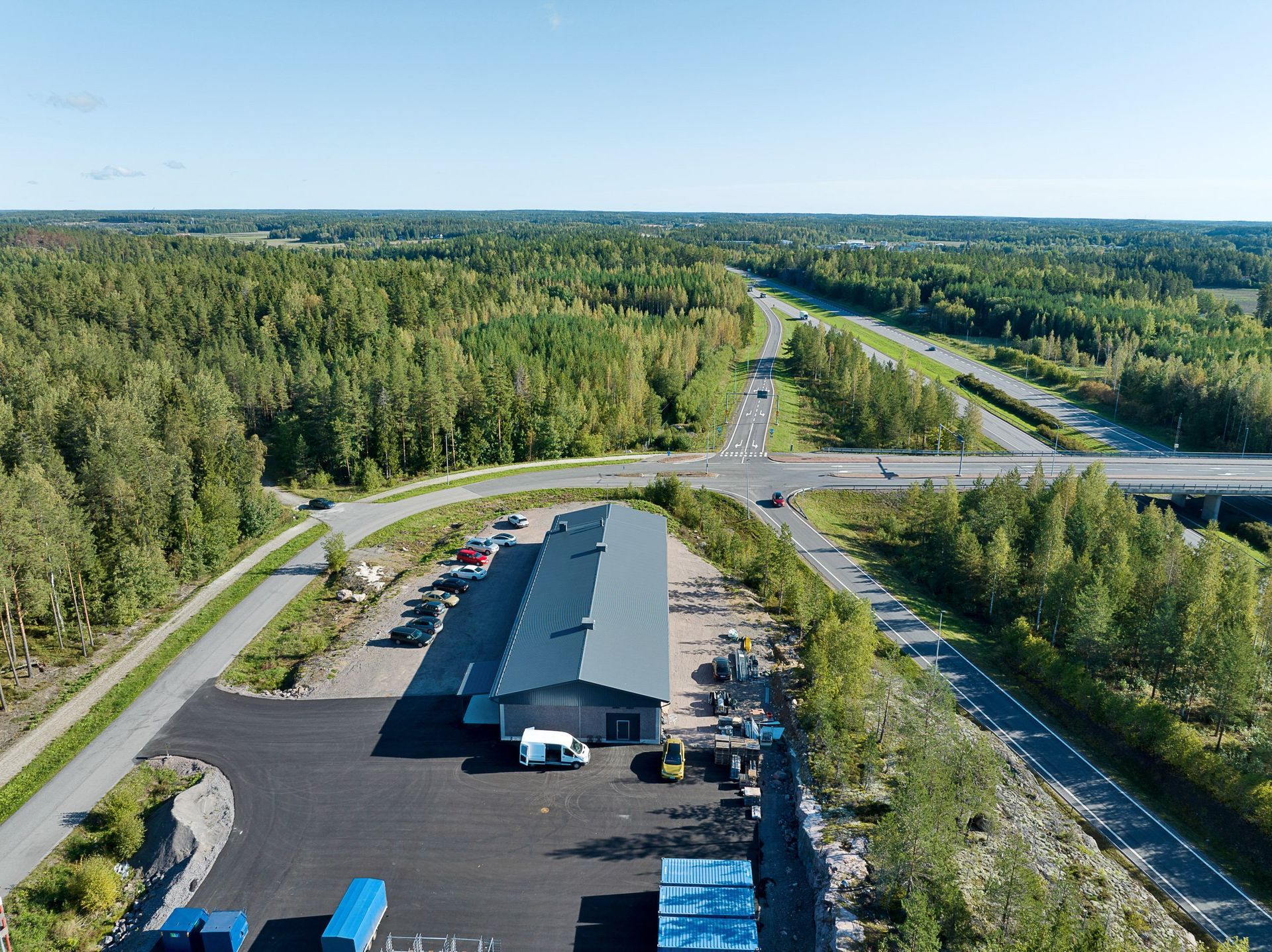 Ilmakuva puiden reunustamasta tienylityspaikasta pitkän harmaakattoisen rakennuksen ja pysäköintialueen lähellä