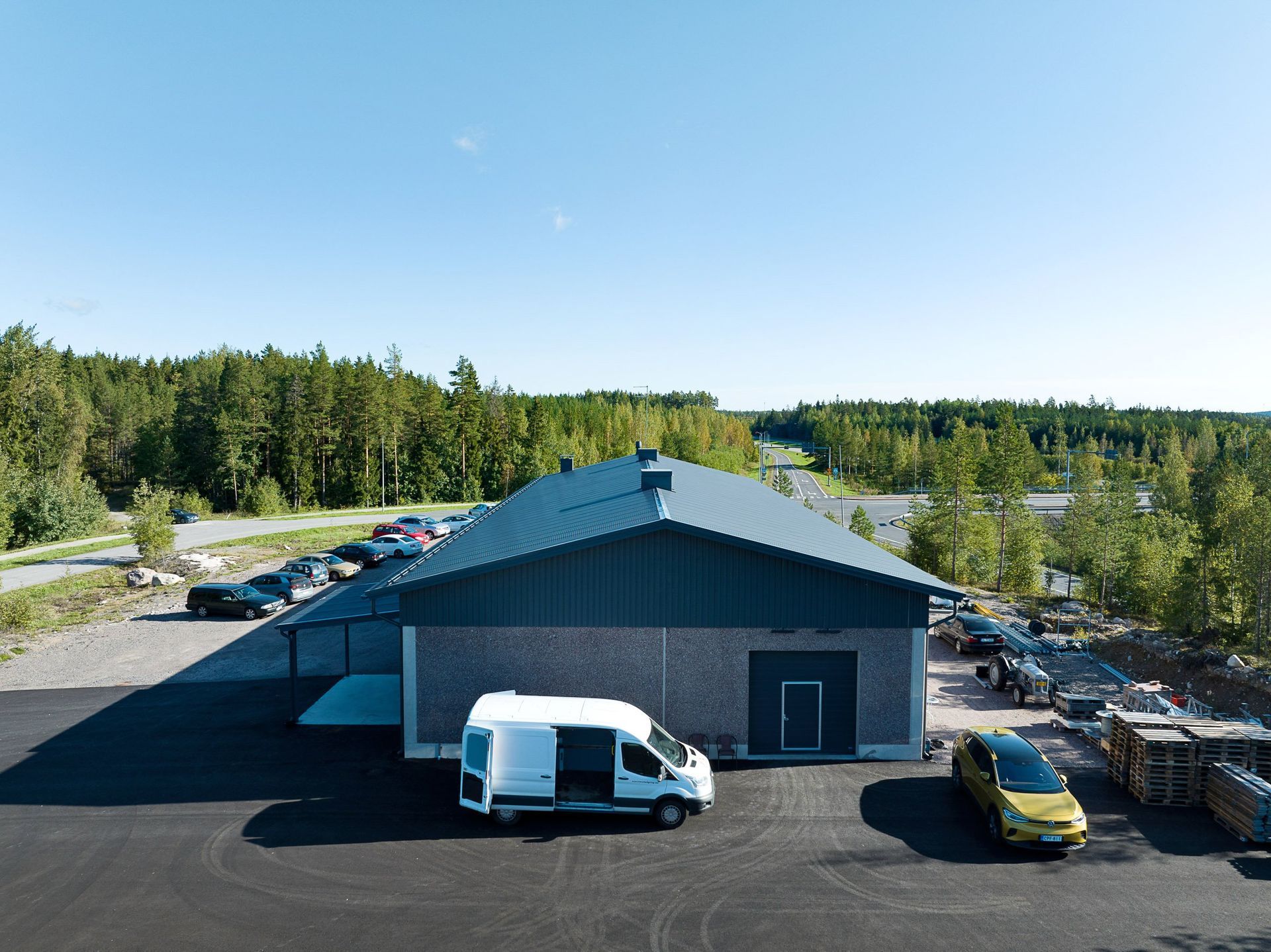 Pieni harmaa teollisuusrakennus, jossa on pysäköity valkoinen pakettiauto, puiden ympäröimänä kirkkaan sinisen taivaan alla