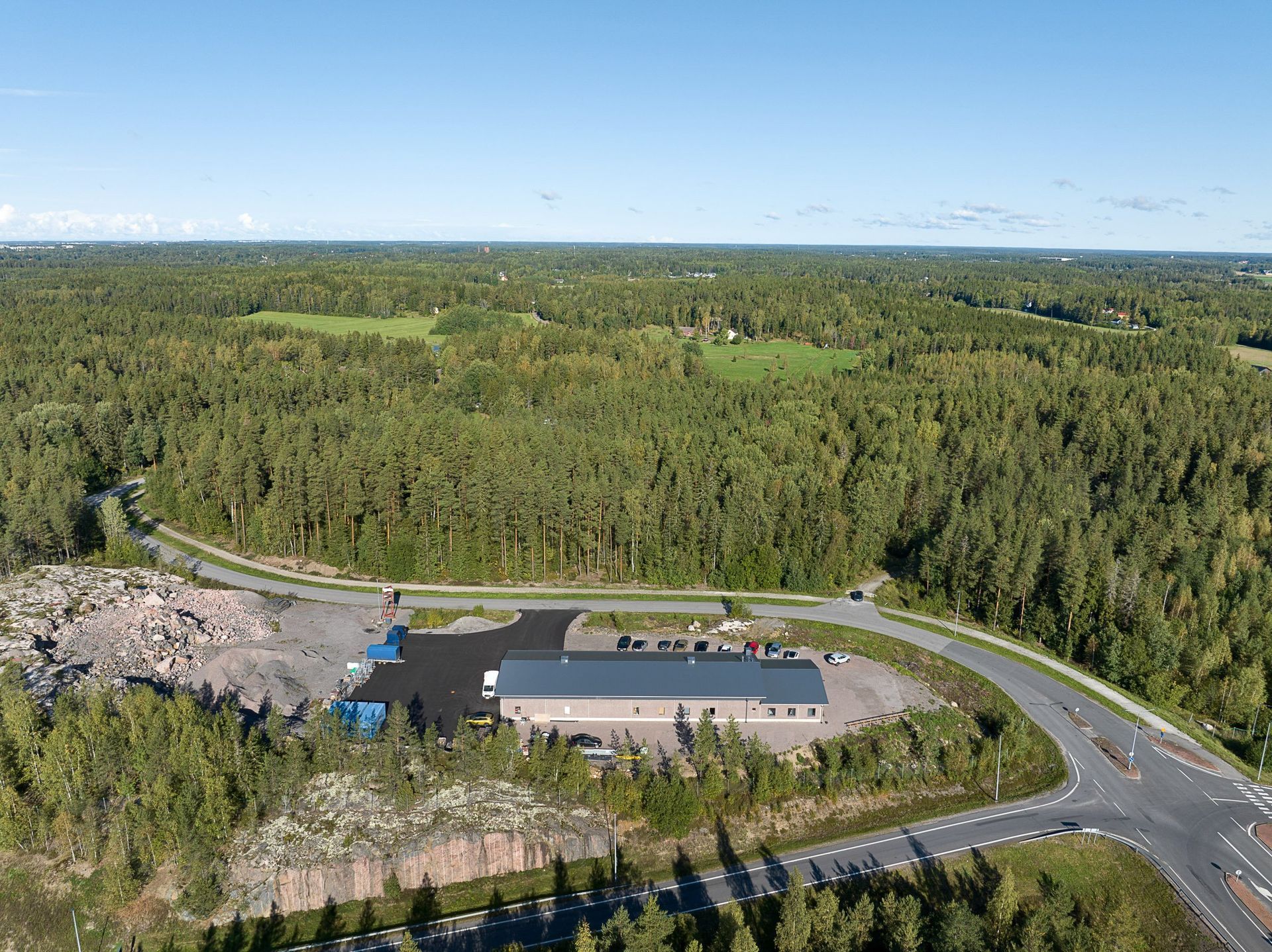 Ilmakuva metsäalueesta, jossa on kaareva tie, rakennus ja pysäköintialue kirkkaan taivaan alla