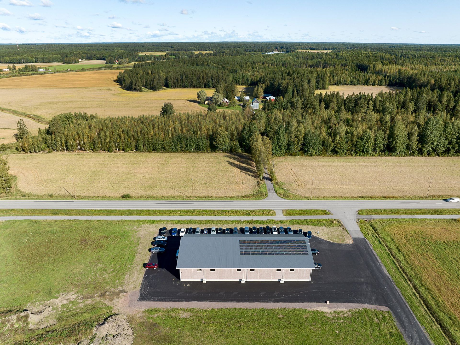 Ilmakuva harmaakattoisesta maaseudun rakennuksesta peltojen ja tiheiden puiden vieressä.