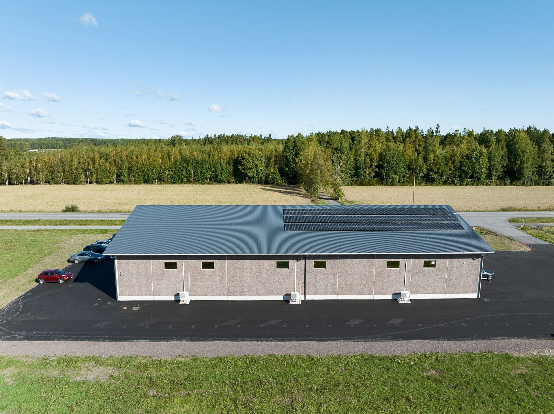 Ilmakuva pienestä tummakattoisesta tiilirakennuksesta tien ja maissipellon vieressä sinisen taivaan alla.