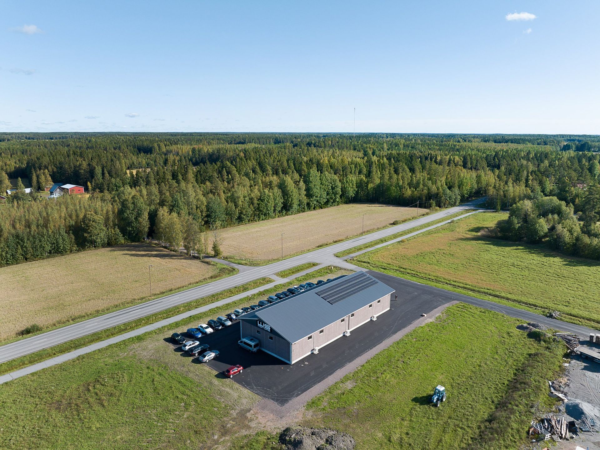 Ilmakuva pitkästä harmaasta rakennuksesta maaseudun pellolla tien vieressä, metsän ympäröimänä.