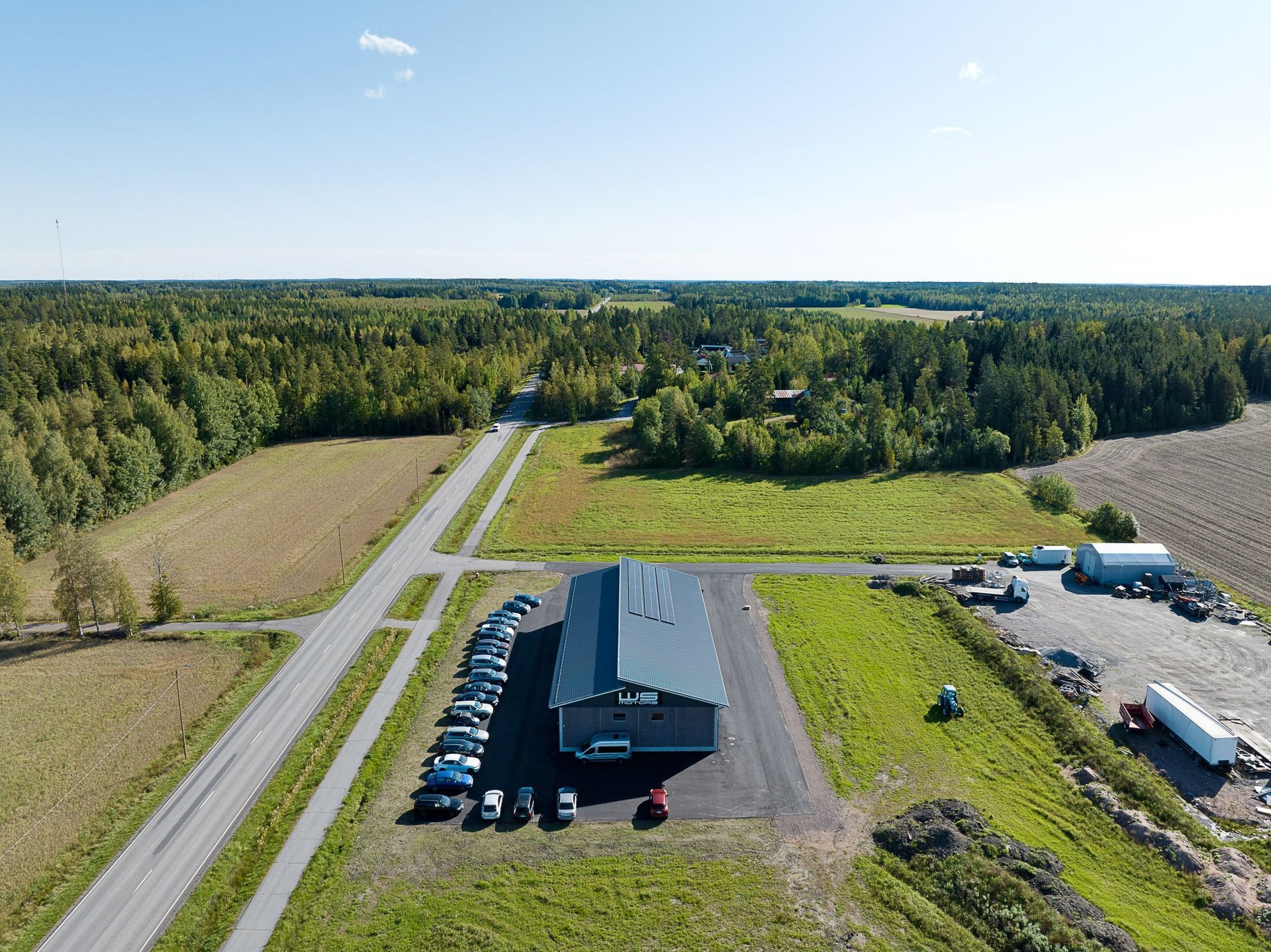Ilmakuva rakennuksesta, jossa on pysäköityjä autoja teiden, peltojen ja tiheän metsän vieressä kirkkaan taivaan alla