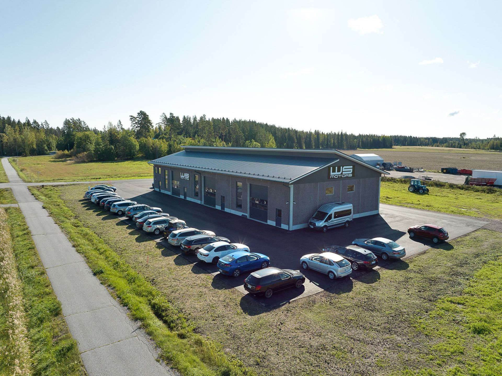 Ilmakuva harmaasta teollisuusrakennuksesta, jossa on paljon pysäköityjä autoja tien varrella olevalla soratietällä.