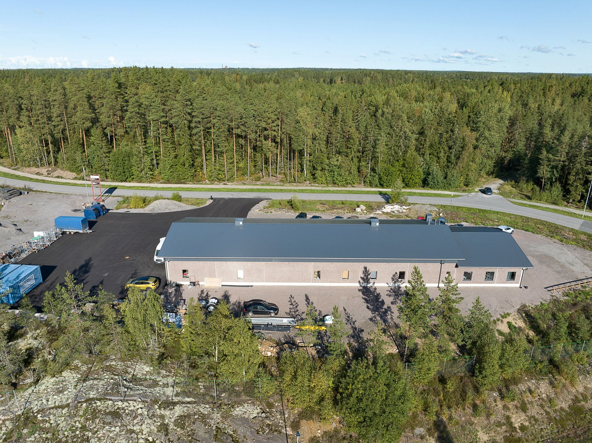 Ilmakuva pitkästä metallikattoisesta rakennuksesta metsäisellä maaseudulla, jossa on pysäköityjä ajoneuvoja ja soratontti