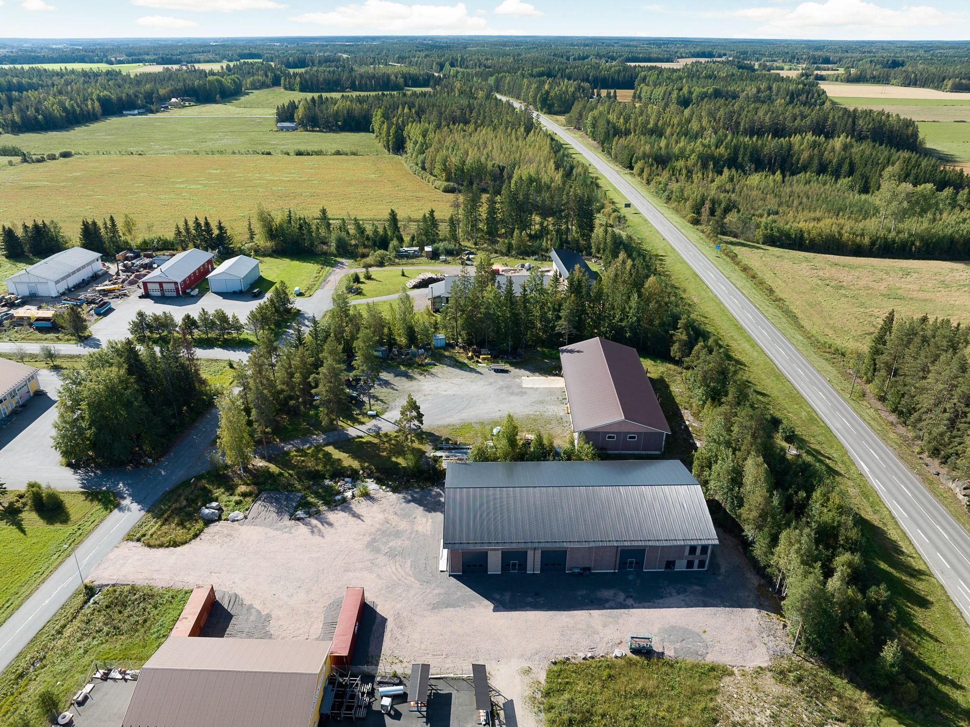 Ilmakuva maaseudun rakennuksista pitkän tien varrella, peltojen ja puiden ympäröimänä