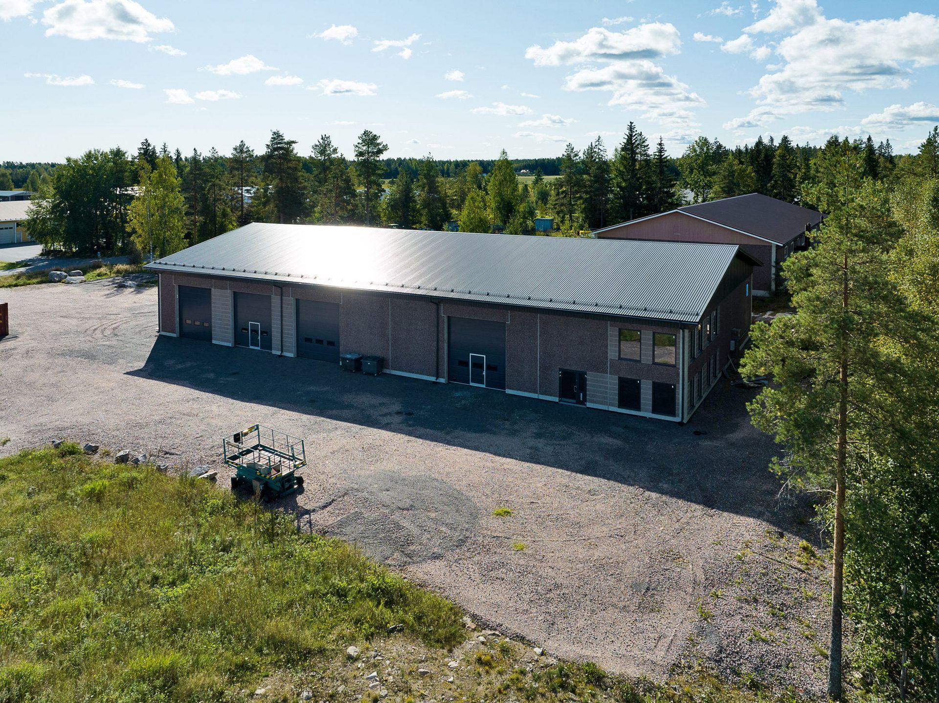 Moderni yksikerroksinen rakennus, jossa on kaareva hopeinen katto, soratontti ja puita kirkkaan taivaan alla