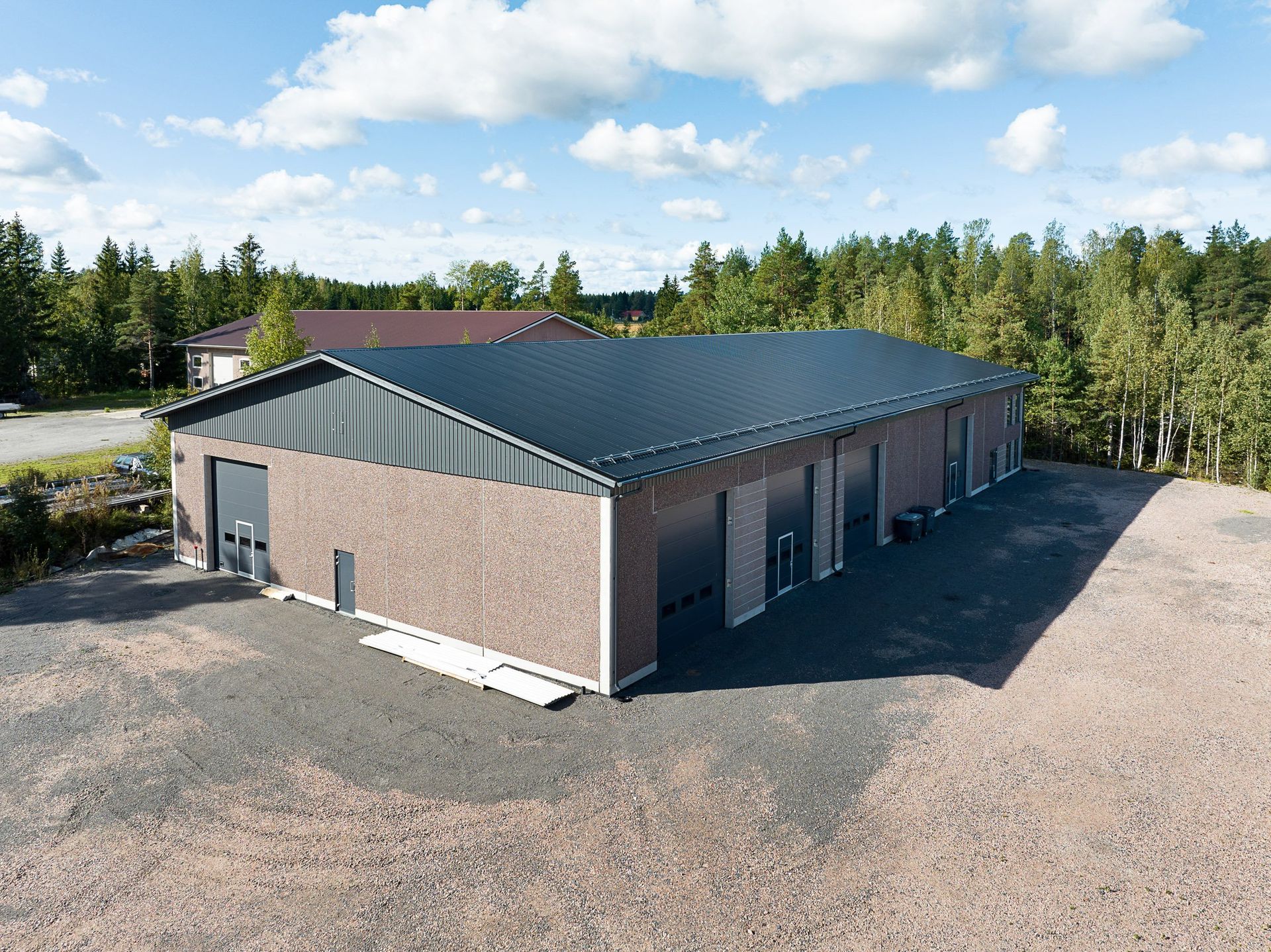 Tiilinen teollisuusrakennus, jossa on tumma metallikatto, soratontilla, puiden ympäröimänä pilvisen taivaan alla