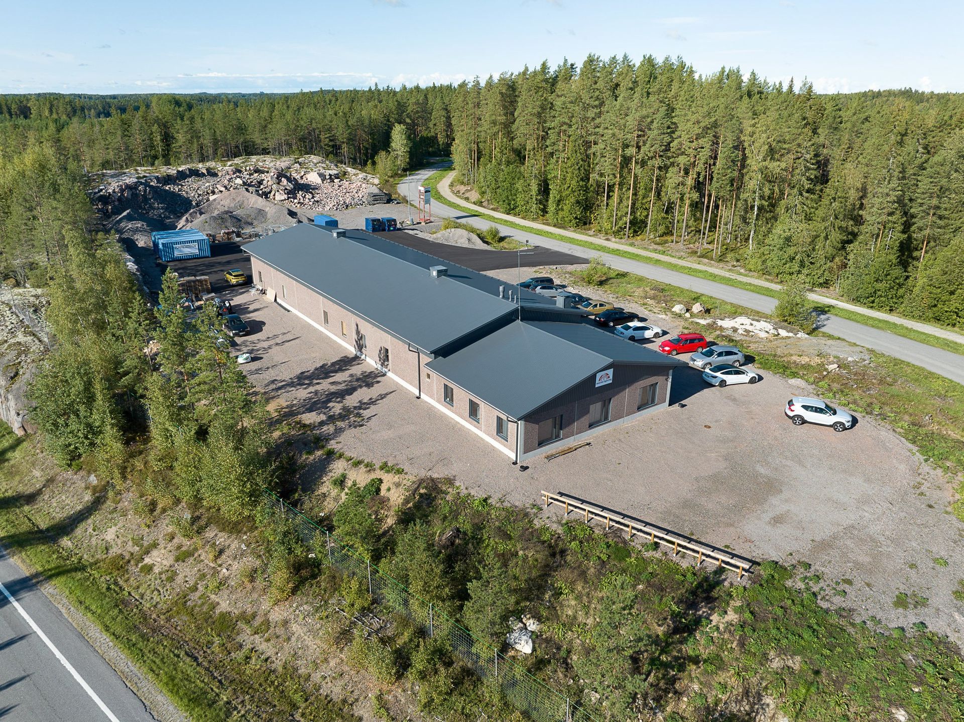 Ilmakuva pitkästä harmaasta rakennuksesta sorakentällä metsätien varrella, jossa on useita pysäköityjä autoja.