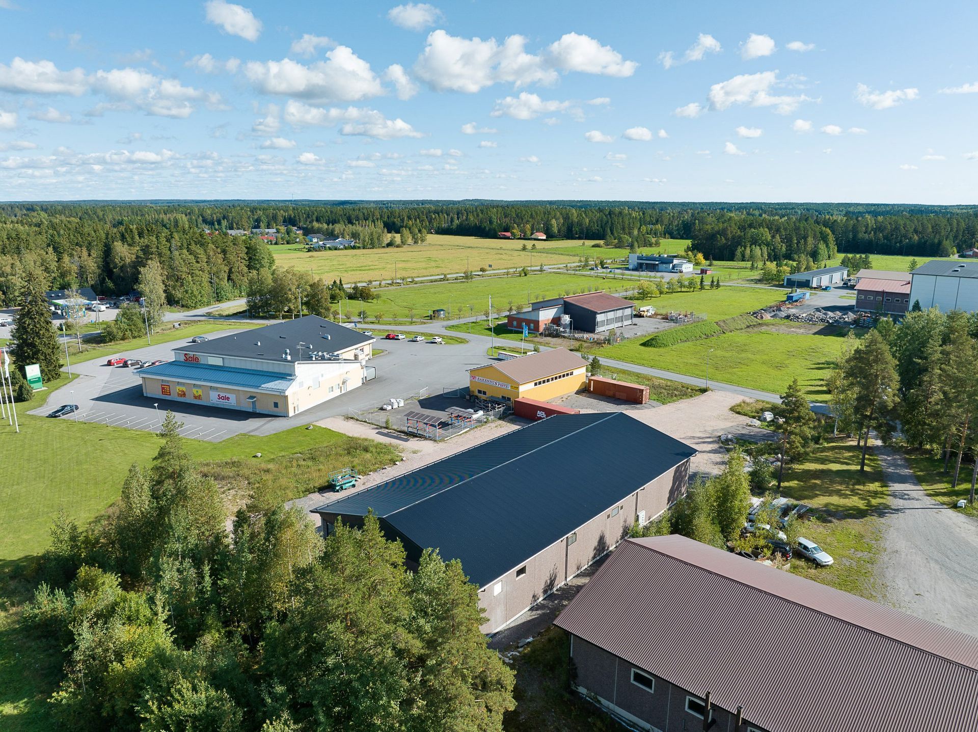 Ilmakuva maaseudun kampukselta, jossa on useita rakennuksia, vihreitä peltoja ja metsä sinisen taivaan alla.