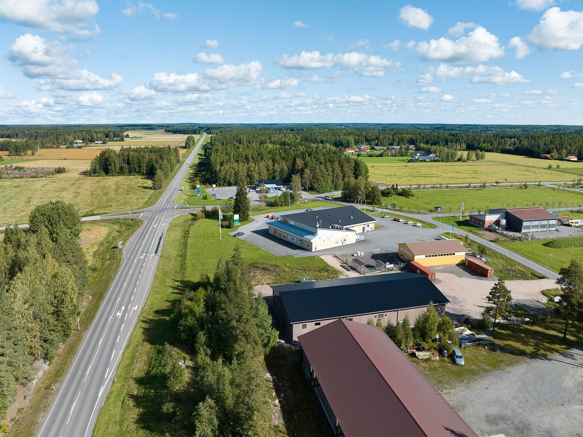 Ilmakuva maaseututieltä peltojen, puiden ja pienten teollisuusrakennusten vieressä osittain pilvisen taivaan alla